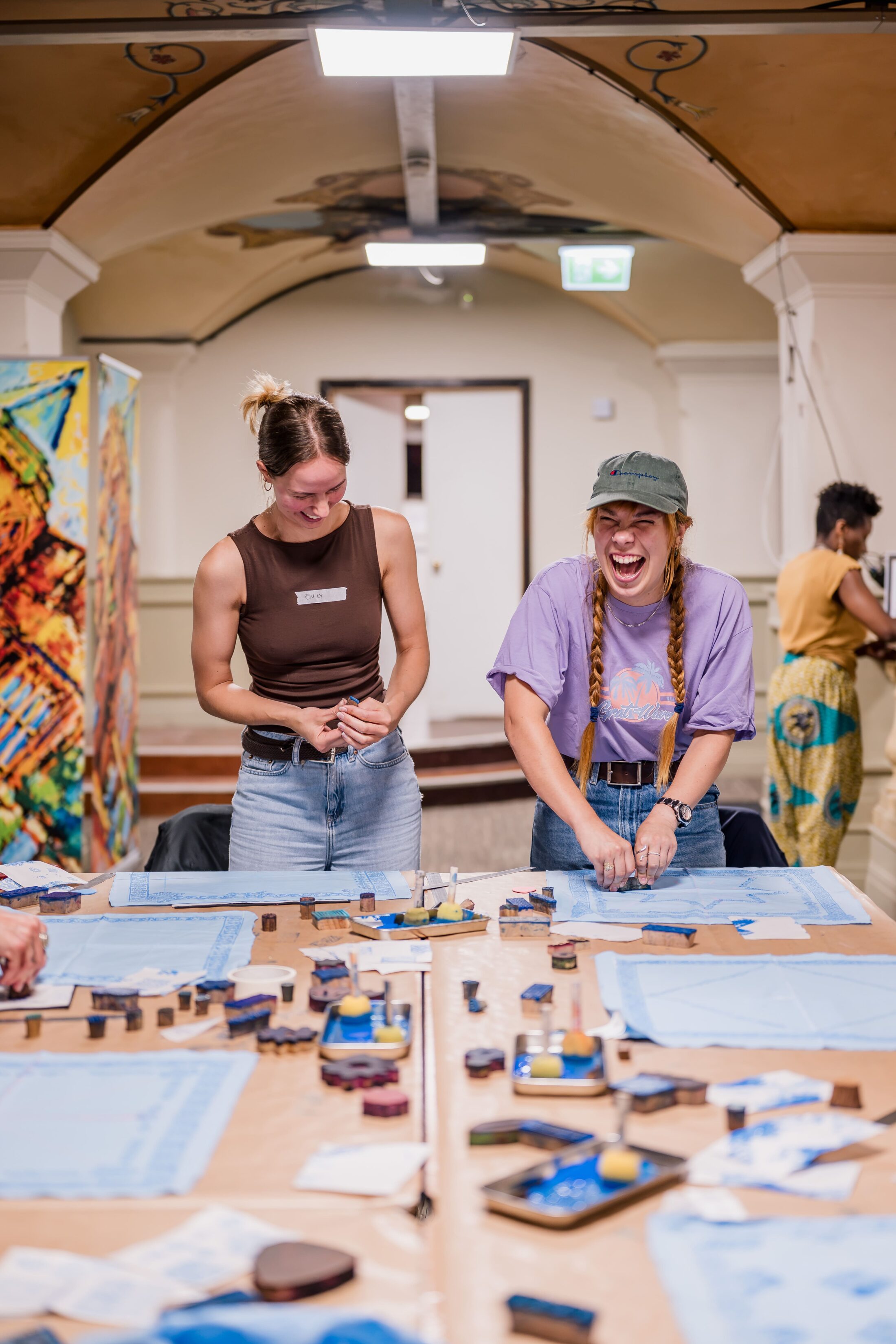 Two young women stand at a table, laughing and working on an art project with blue fabric and stamps in a brightly lit room. Other people are also participating in the activity in the background.