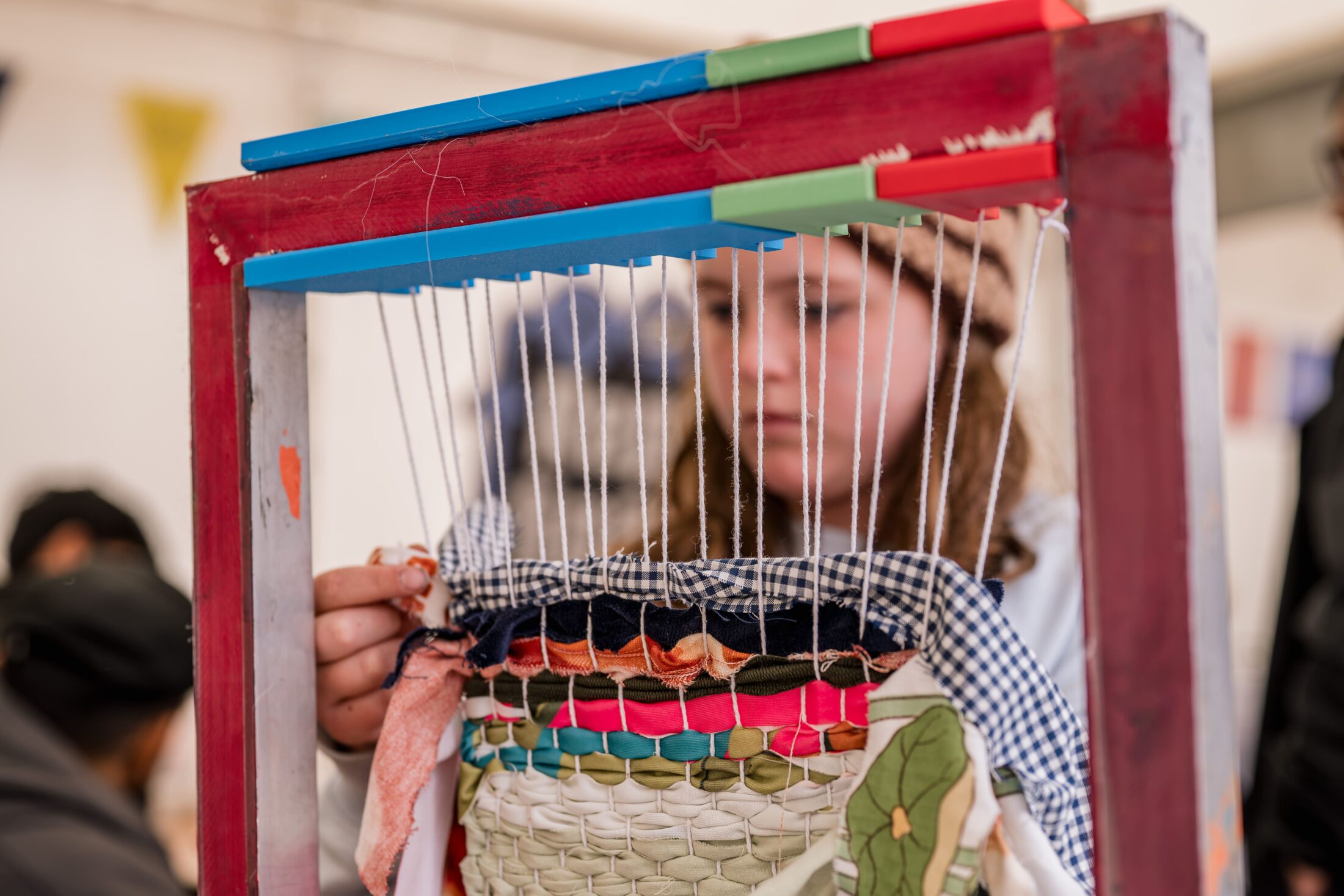 A person weaves colourful fabric strips on a loom, focusing intently on their craft. The loom has red, blue, and green parts, and woven textiles with various colours and textures are visible.