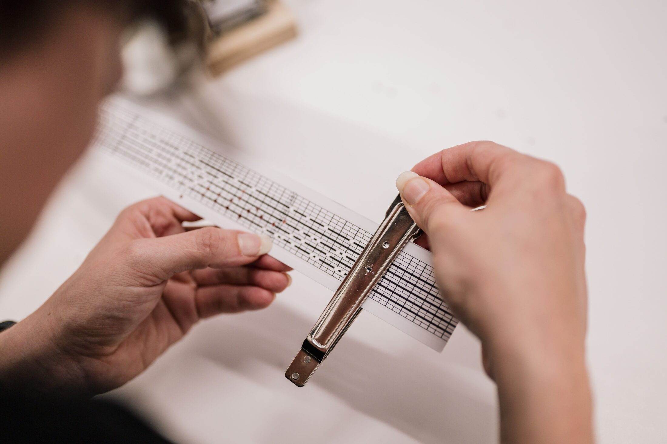 A person uses a metal stapler to attach a sheet of paper with musical notes printed on it. The background is white, and the scene focuses on the hands and the paper.
