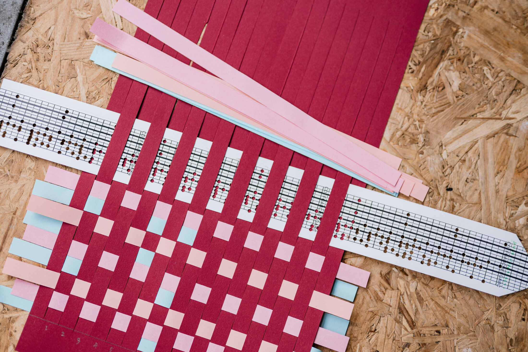 Red paper with slits is woven with pink, blue, and white strips, some of which have music notes printed on them. The woven paper is placed on a wooden surface.