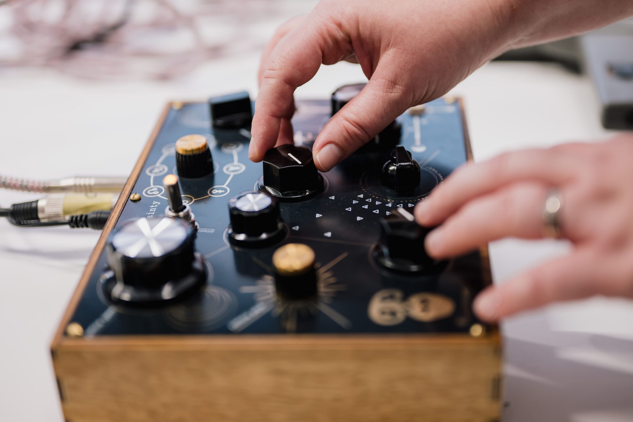 Two hands adjust knobs on a black audio control box with various dials, gold details, and connected cables, suggesting music or sound manipulation.