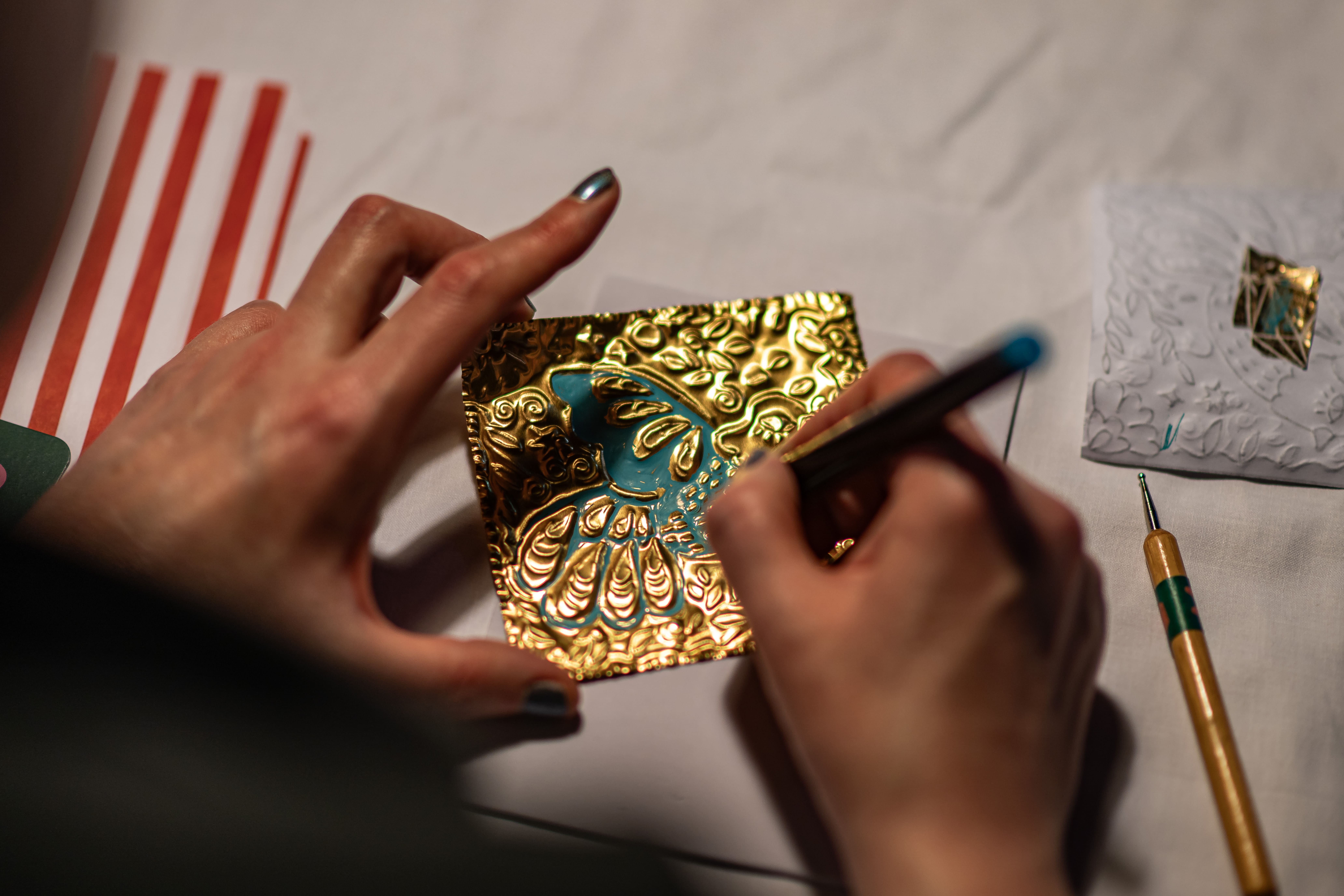 A person uses a stylus to emboss a decorative gold metal sheet with a blue centre, creating intricate patterns. Various tools and materials are visible on a white surface nearby.