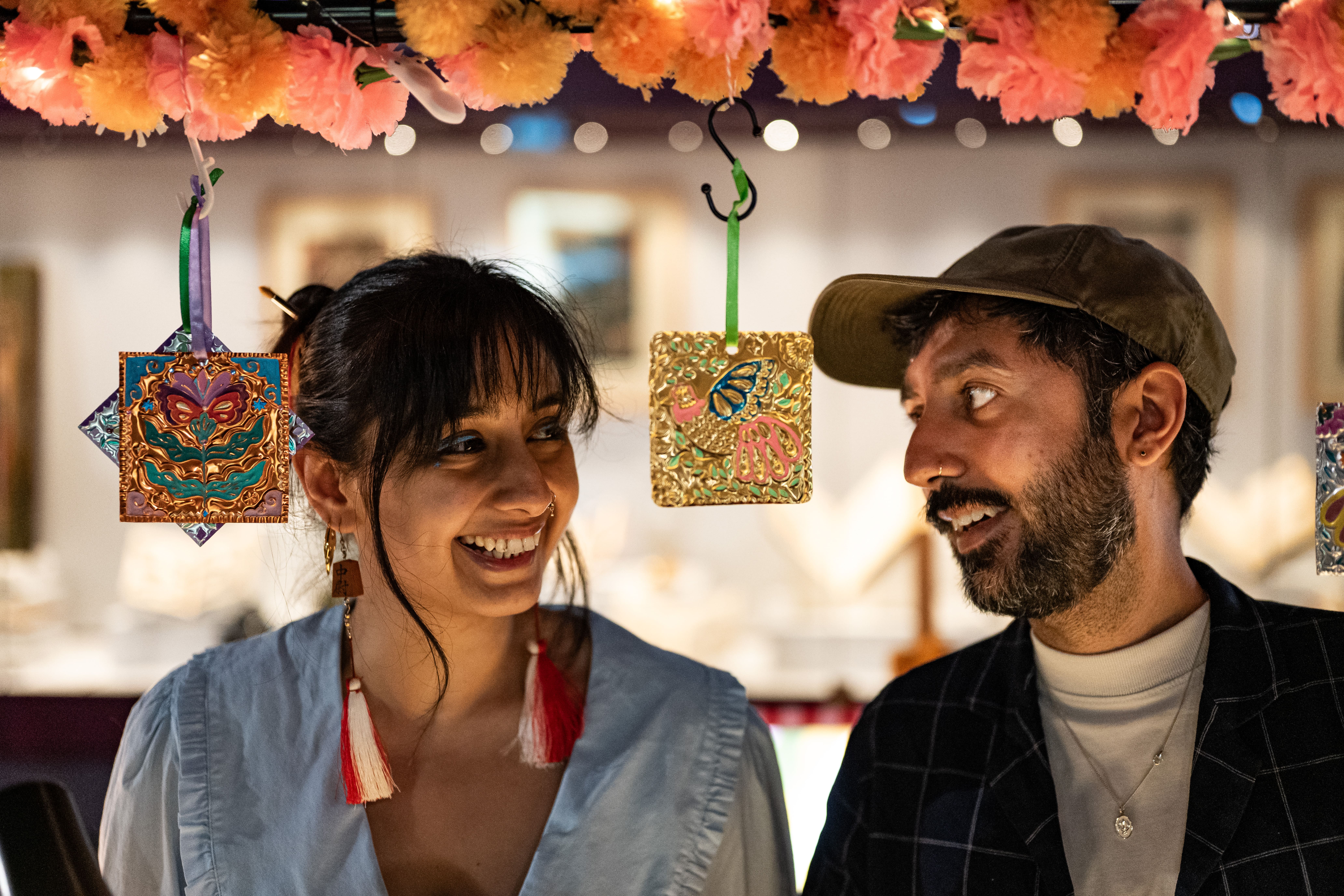 Two people smile at each other under a row of colourful hanging decorations with flowers above. The background is softly blurred, highlighting their joyful expressions and festive atmosphere.