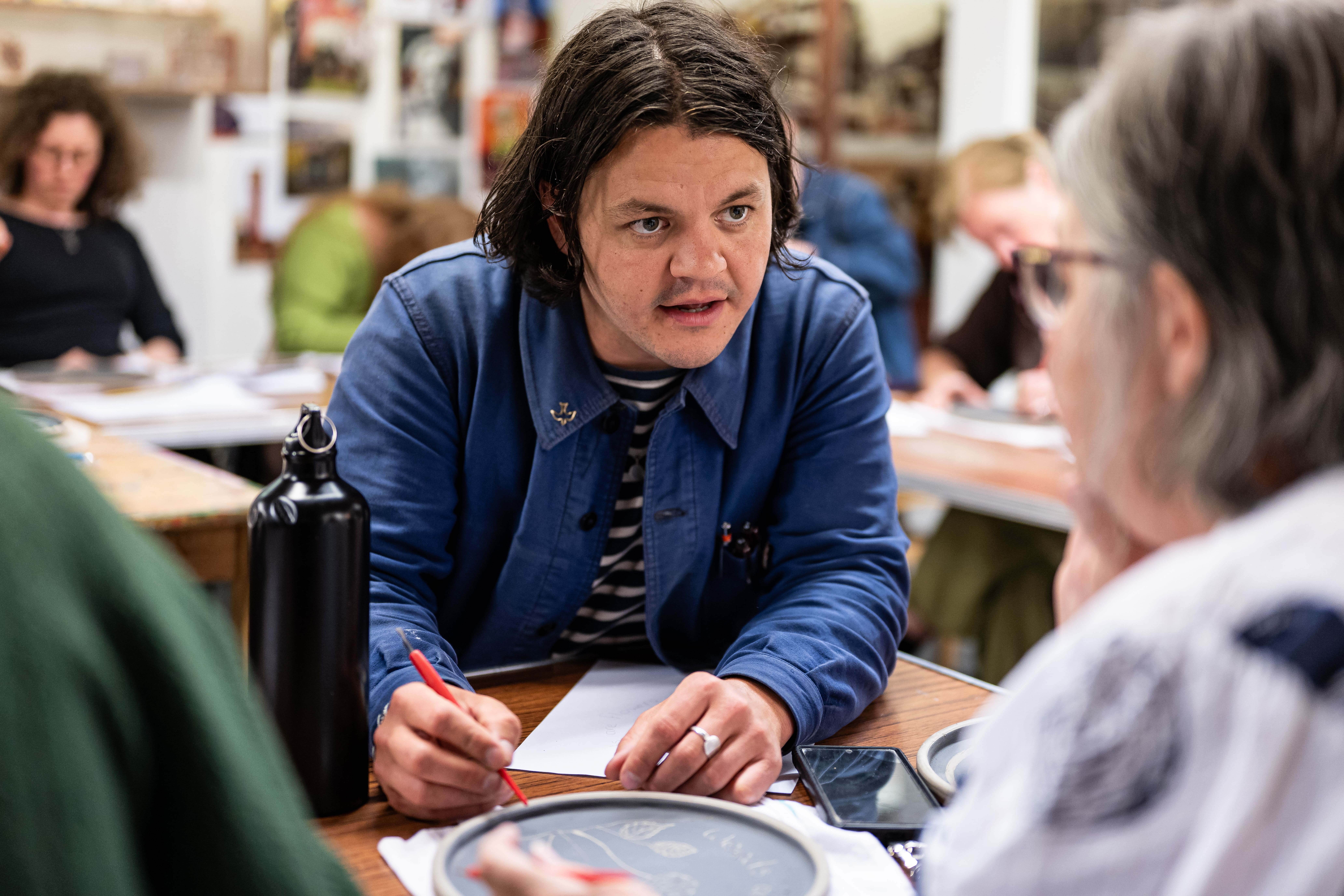 A person with shoulder-length dark hair speaks to another person in a classroom, holding a pencil and paper. Other people are seated and writing in the background.