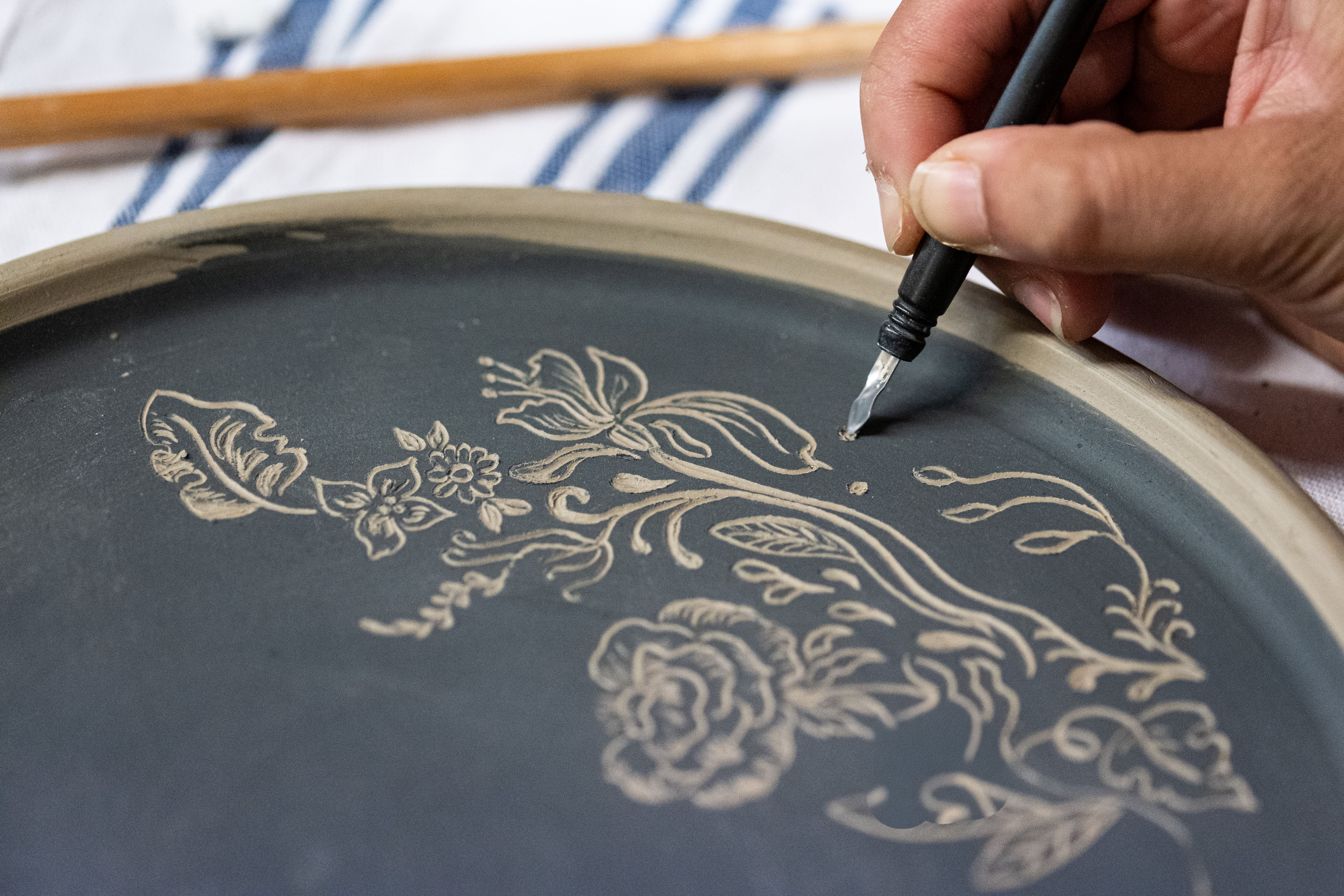 A hand uses a pointed tool to carve intricate floral designs into a dark clay plate, with wooden tools and a striped cloth in the background.