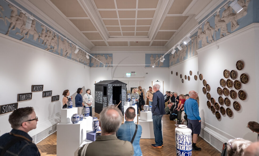A group of people observe pottery and wall art in a bright, ornate gallery space; some listen to a speaker, whilst others view the exhibits.