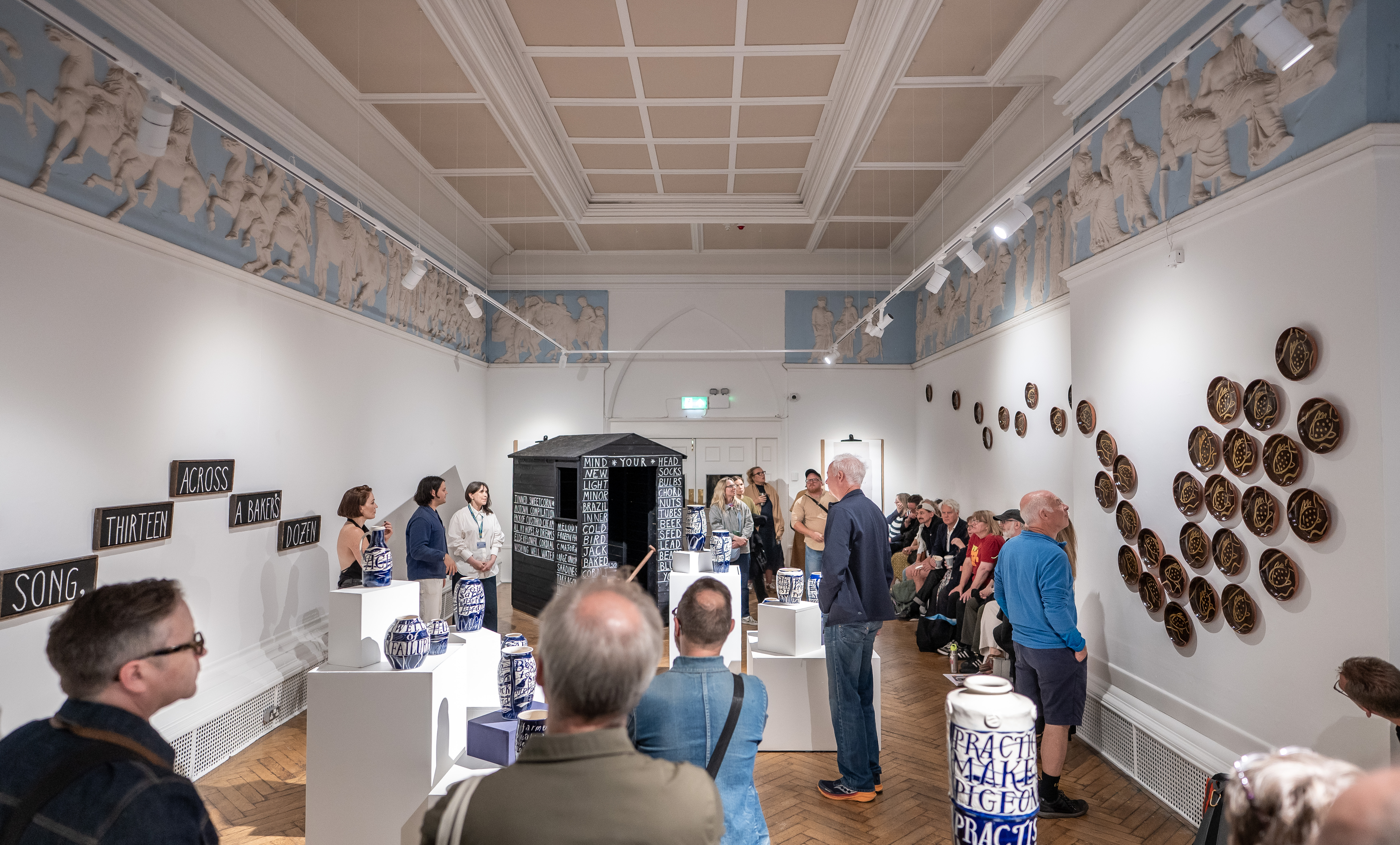 A group of people observe pottery and wall art in a bright, ornate gallery space; some listen to a speaker, whilst others view the exhibits.