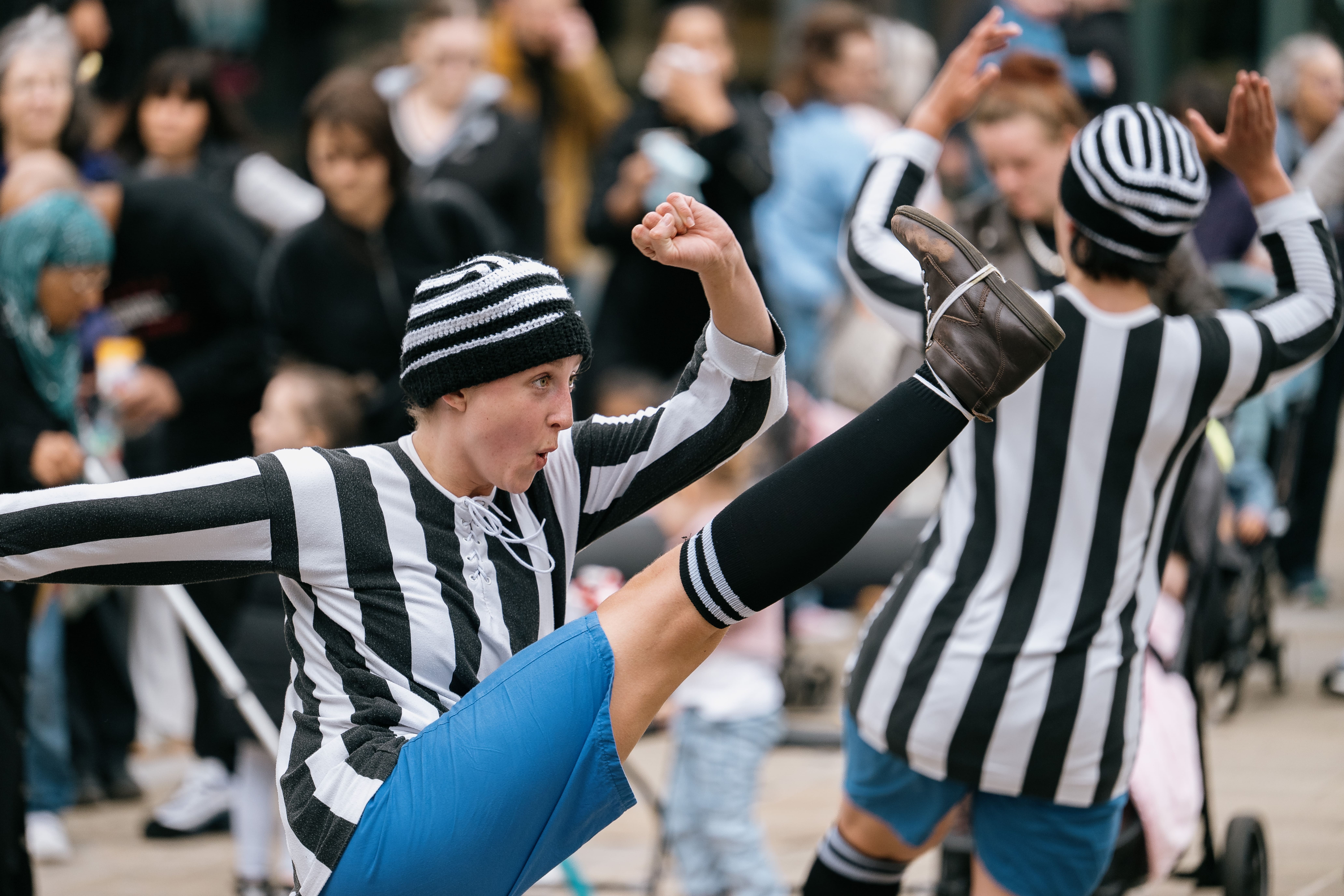 Two performers in black-and-white striped shirts, blue shorts, and striped hats energetically kick their legs high during a lively outdoor event, with a crowd of people watching in the background.