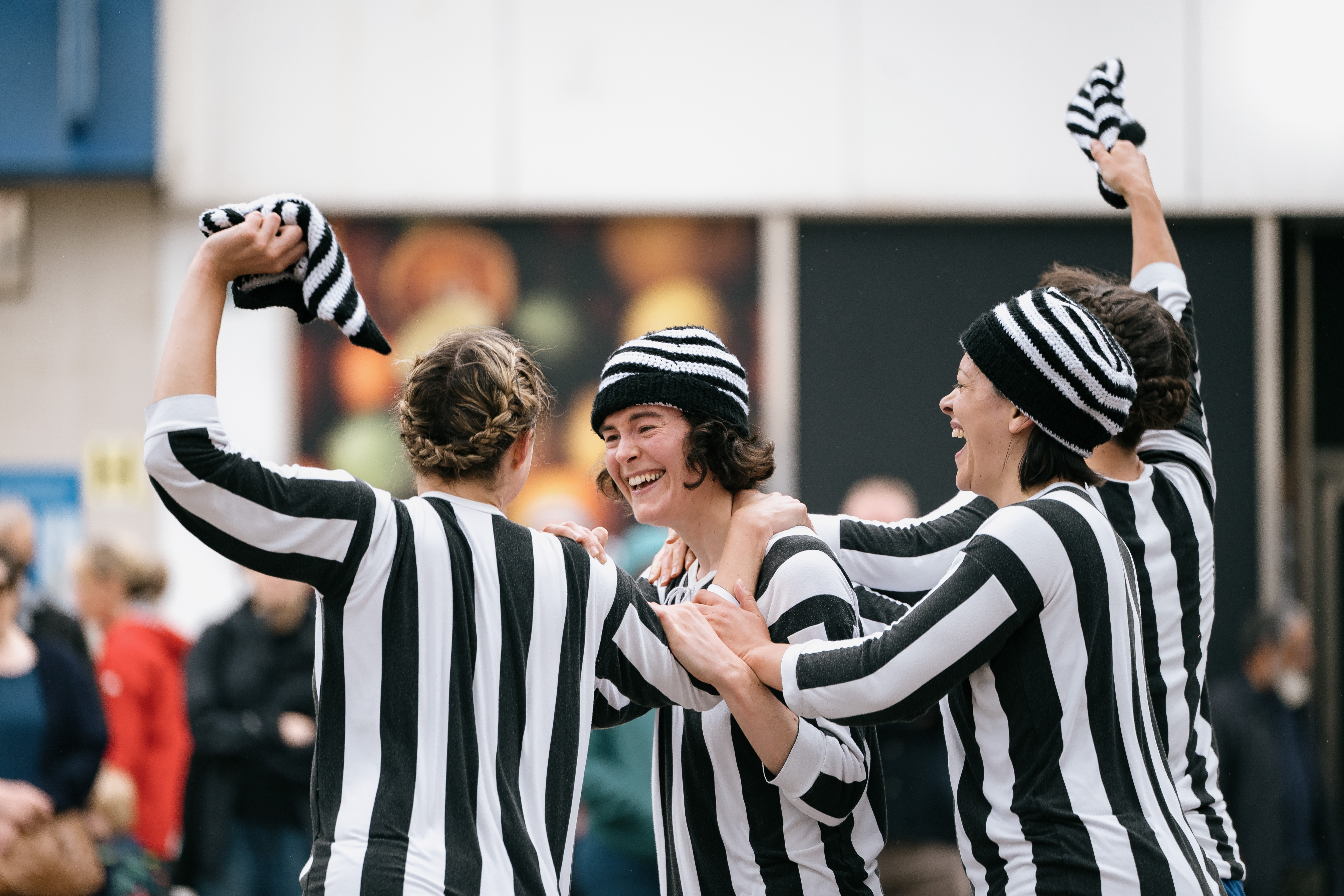 Four people in black and white striped shirts and hats celebrate together outdoors, smiling and waving their hats, with a blurred crowd and colourful lights in the background.