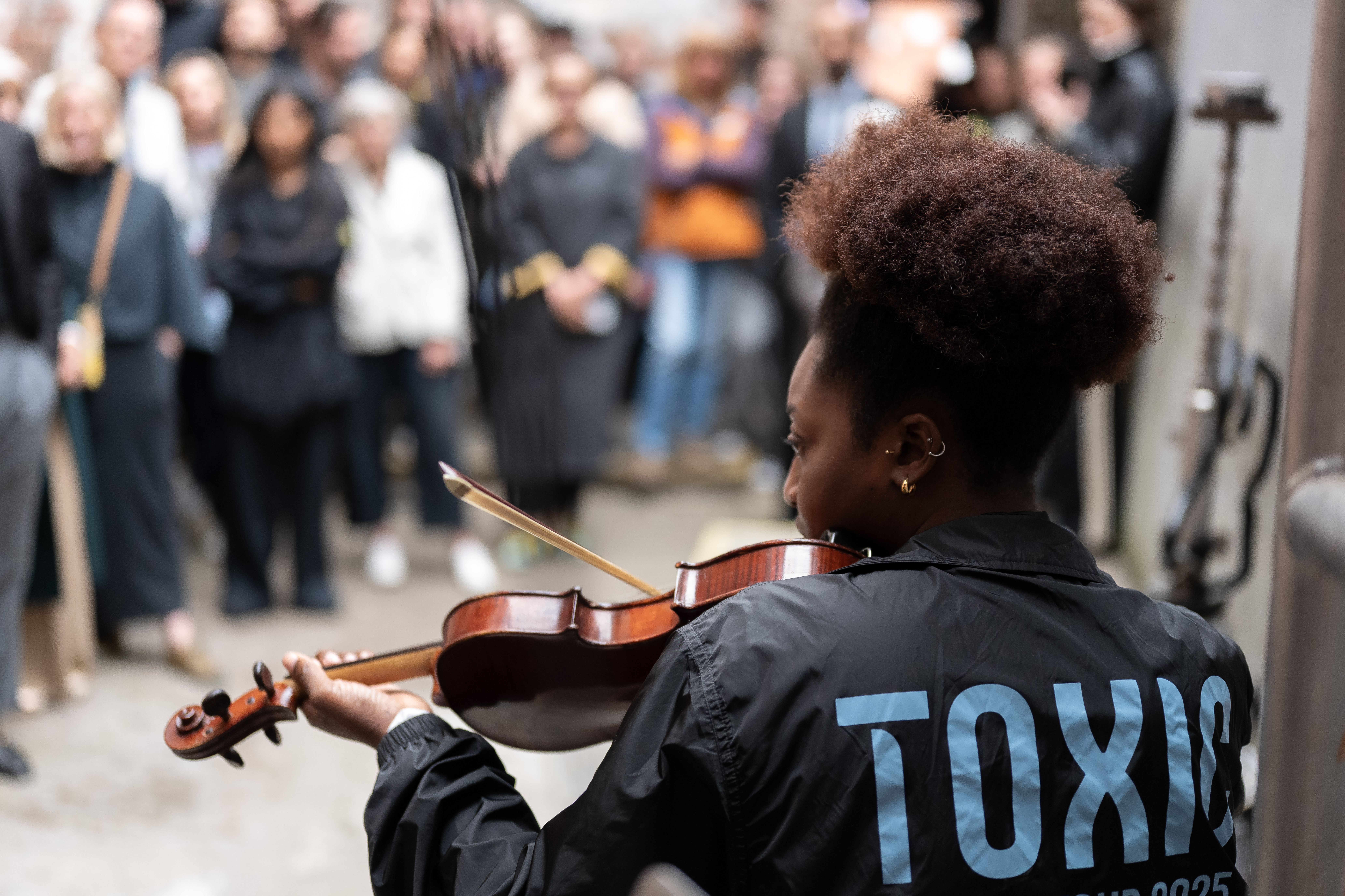 A musician wearing a jacket with TOXIC on the back plays the violin outdoors, whilst a blurred crowd watches in the background.