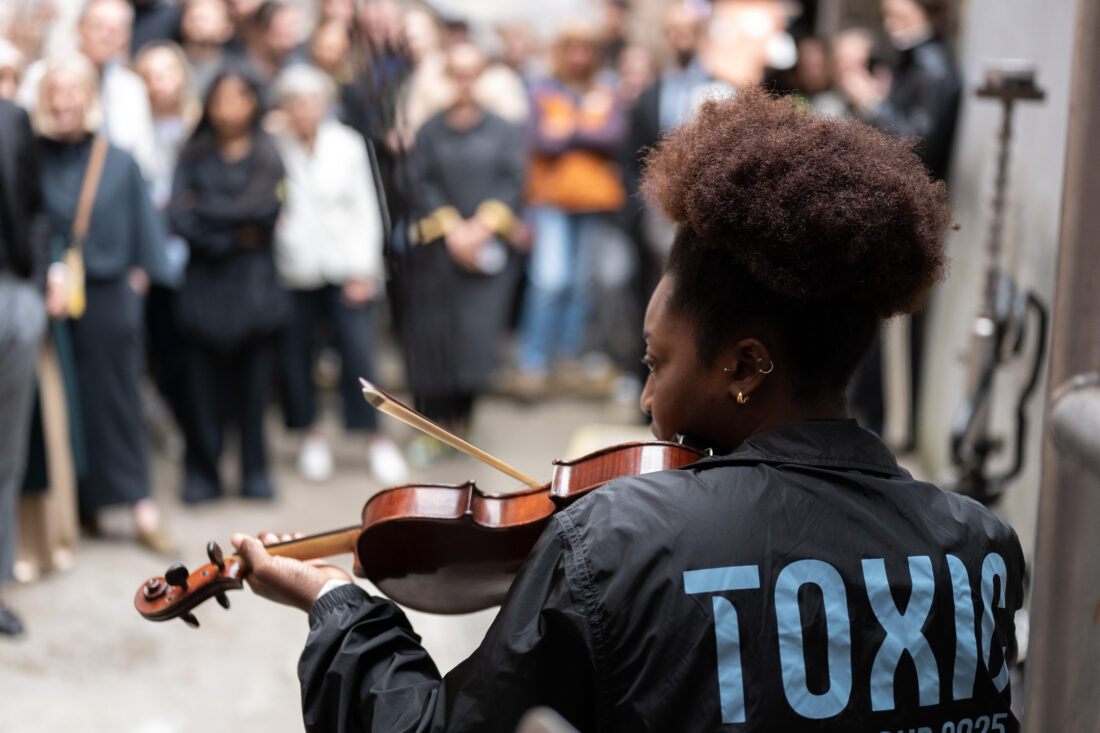 A violinist with a large afro plays for a blurred crowd. She wears a black jacket with TOXIC in large blue letters on the back.