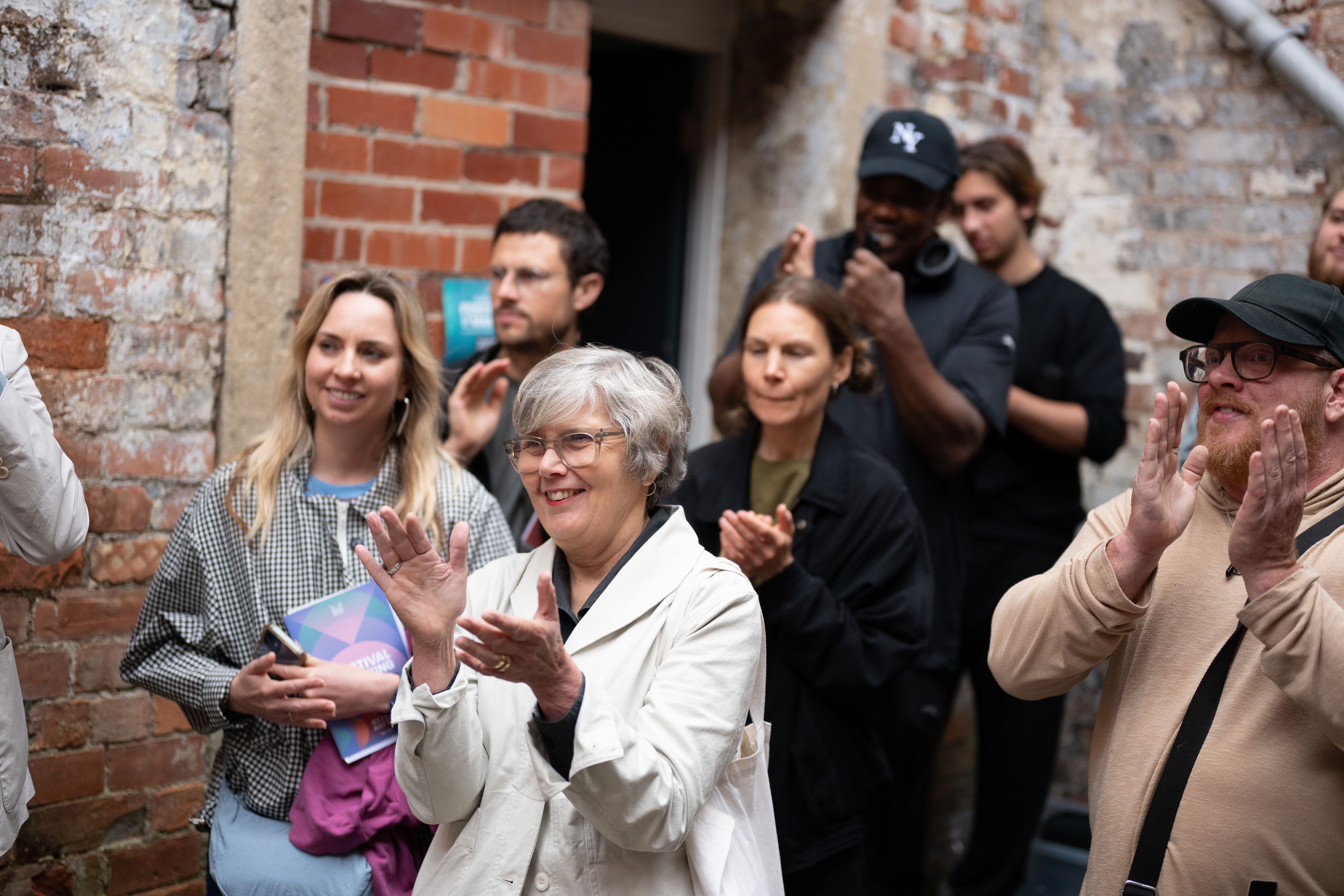 A diverse group of people stand in front of a brick wall, smiling and applauding. Some hold notebooks and wear jackets, creating a casual and friendly atmosphere.