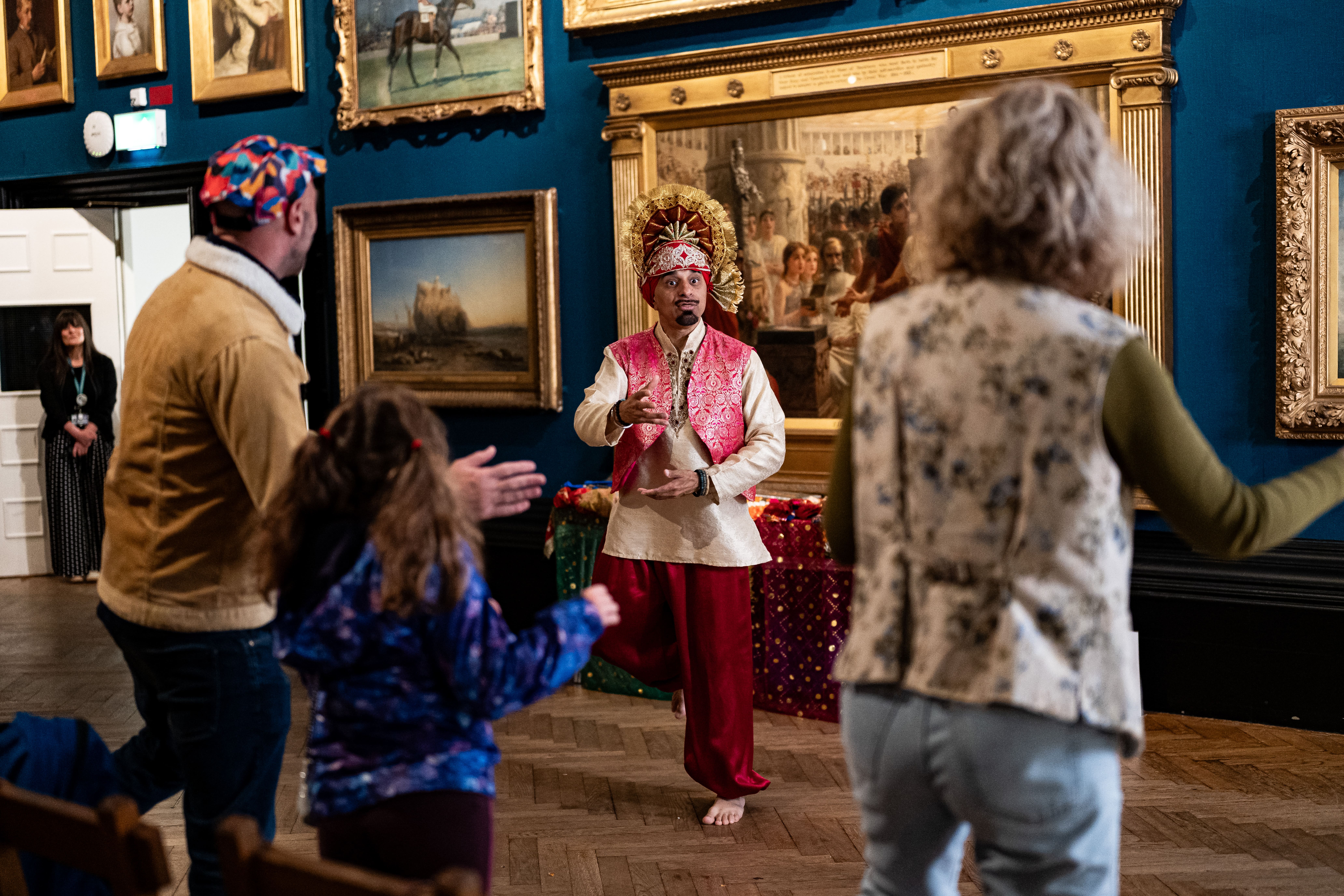A performer in a colourful traditional outfit dances in an art gallery, while a small audience watches and joins in. Framed paintings line the dark blue walls of the gallery.