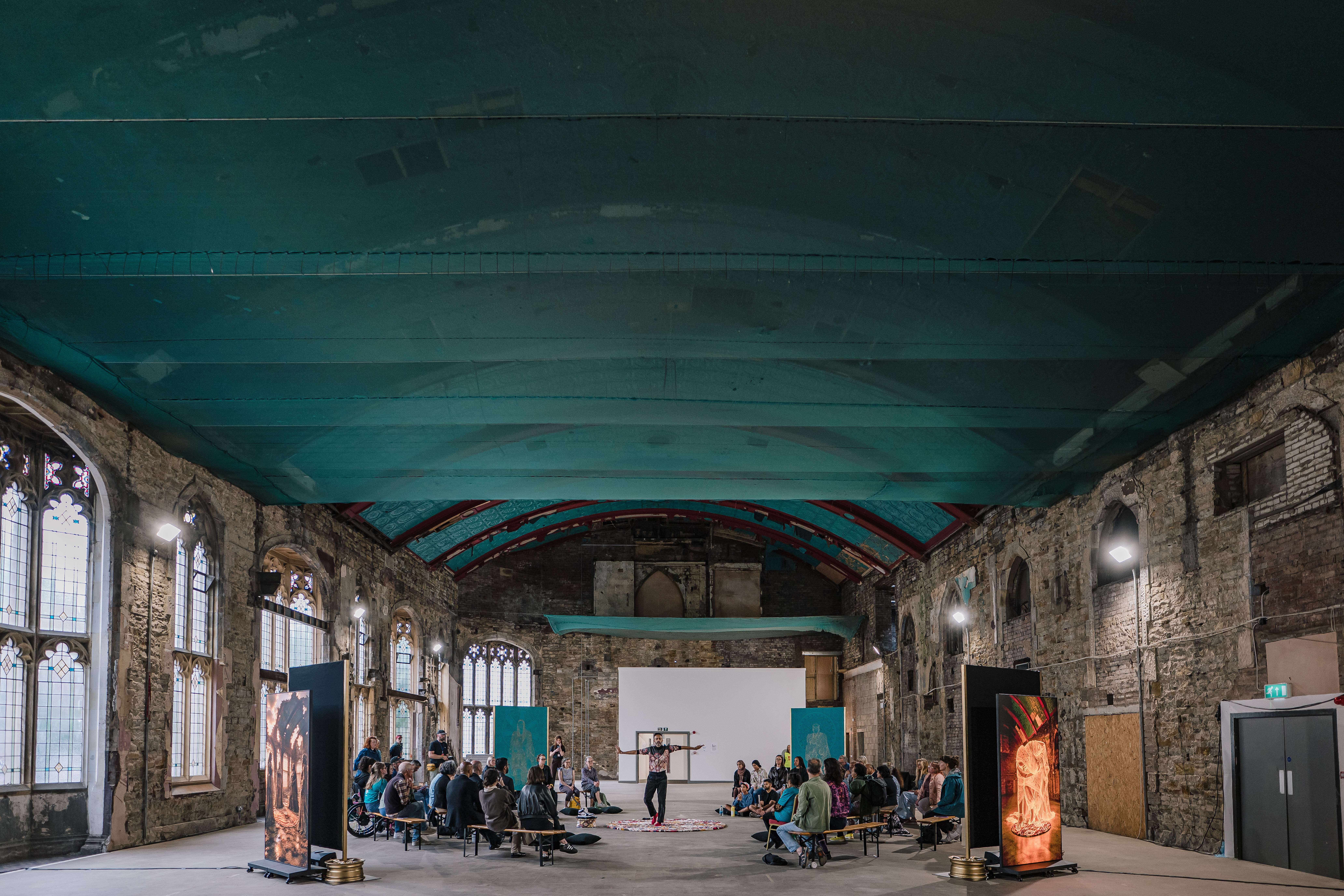 A group of people sit on benches in a large, old hall with high arched windows, exposed brick walls, and a turquoise ceiling, facing a performer in the centre.