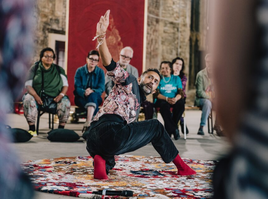 A person in patterned clothes and red socks performs a dance on a colourful rug while a seated audience watches in an indoor setting.