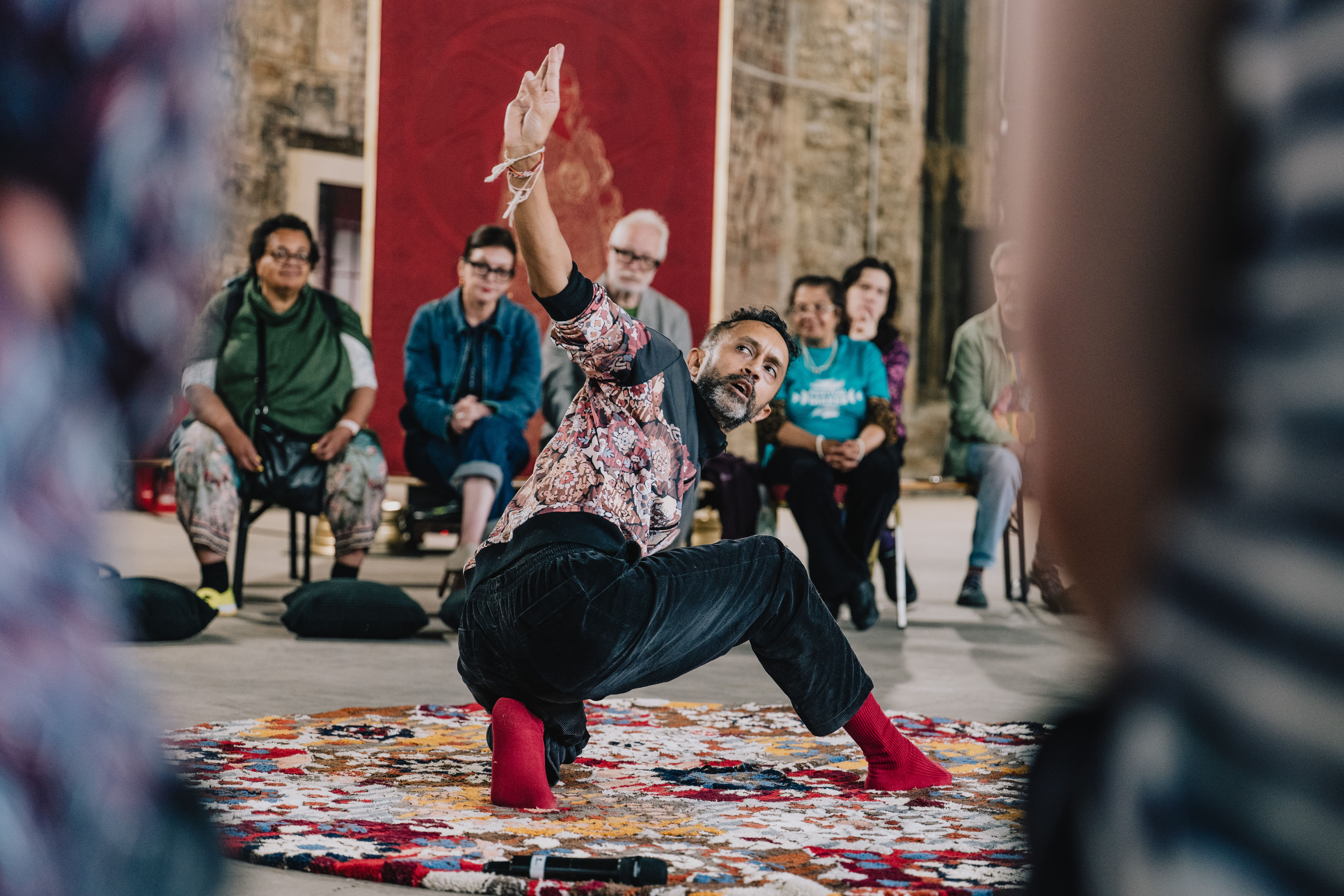 A person in patterned clothes and red socks performs a dance on a colourful rug while a seated audience watches in an indoor setting.