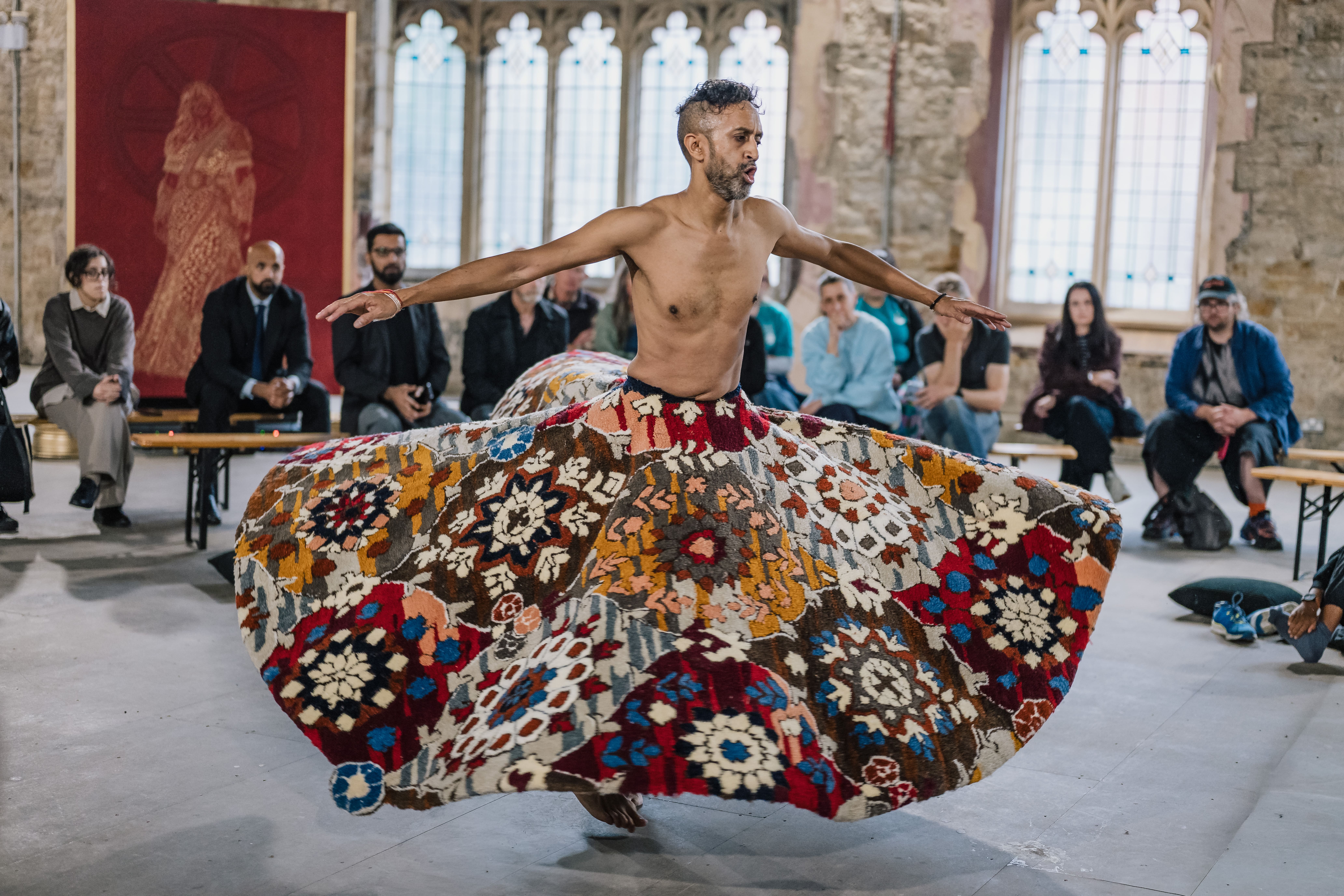 A barefoot, shirtless dancer spins in a colourful, patterned skirt inside a stone hall, while seated people watch against tall, arched windows.