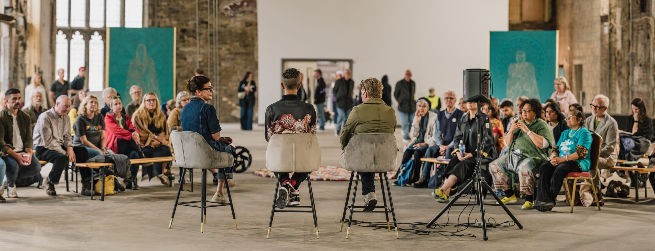Three people sit on chairs facing an audience in a spacious, industrial-style room. The audience watches attentively. Large turquoise art panels are displayed in the background.