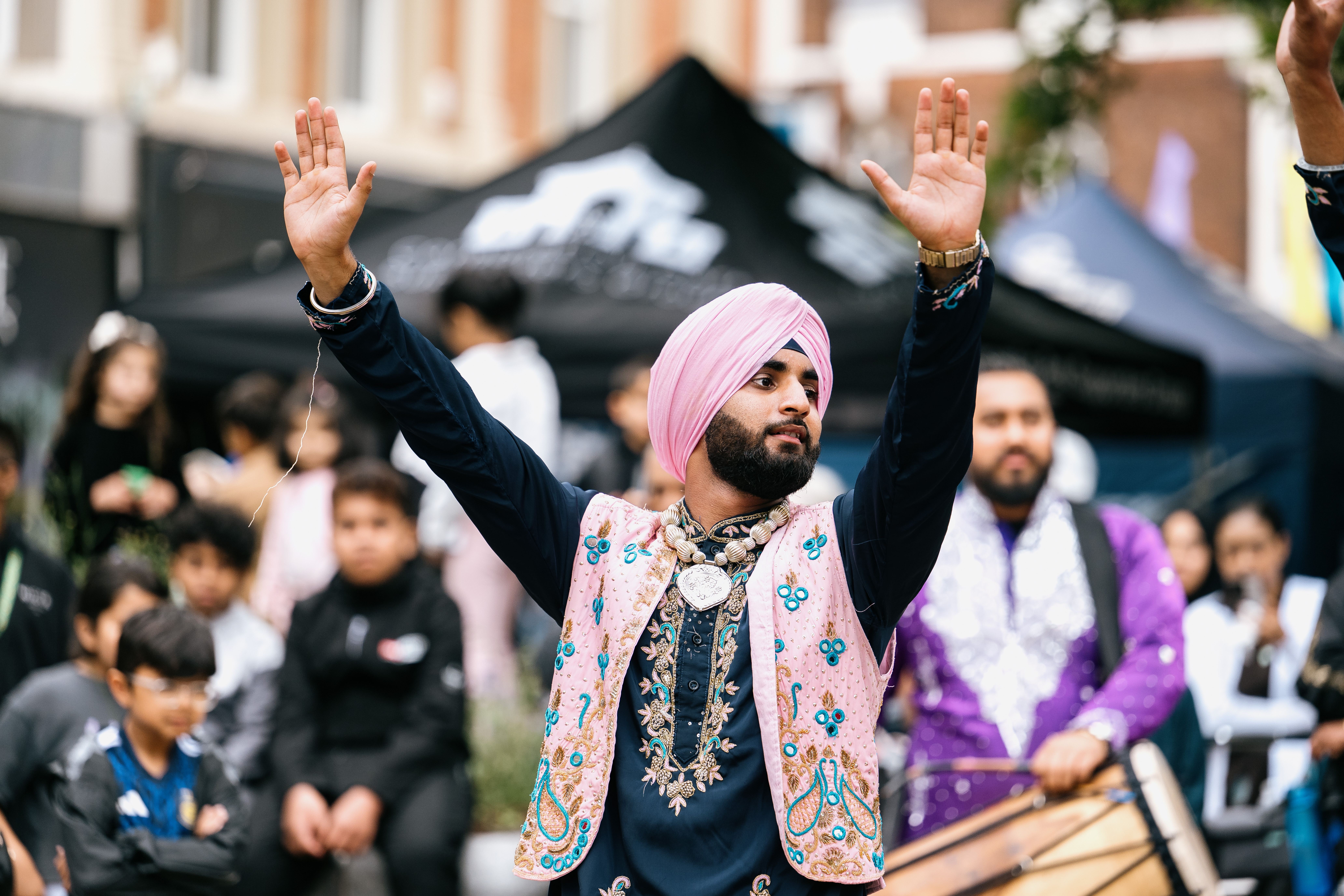 A man in a pink turban and embroidered waistcoat dances with raised arms at an outdoor event, surrounded by people and tents in the background.