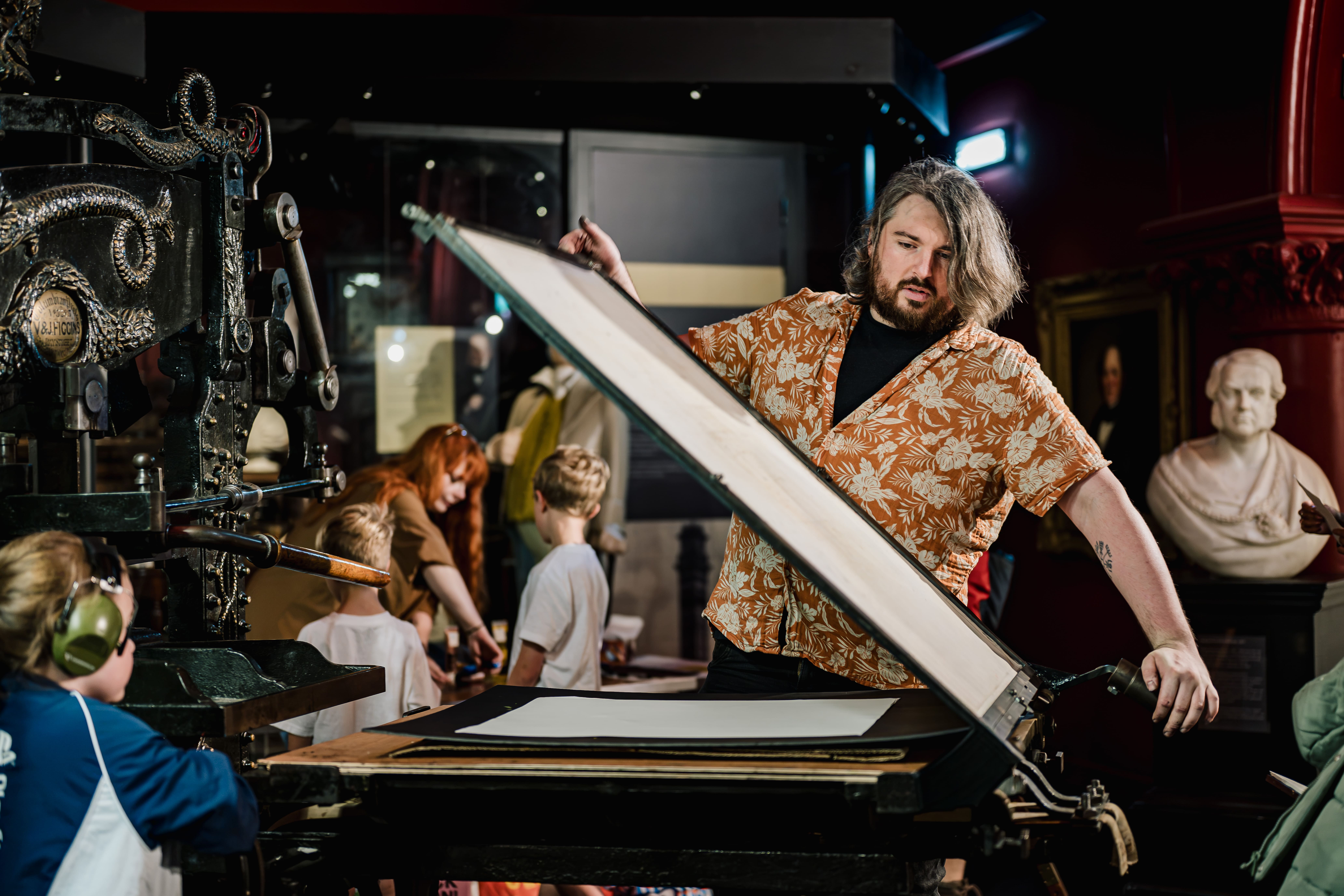 A person in a patterned shirt operates a large vintage printing press, whilst children and adults observe in a museum setting with statues in the background.
