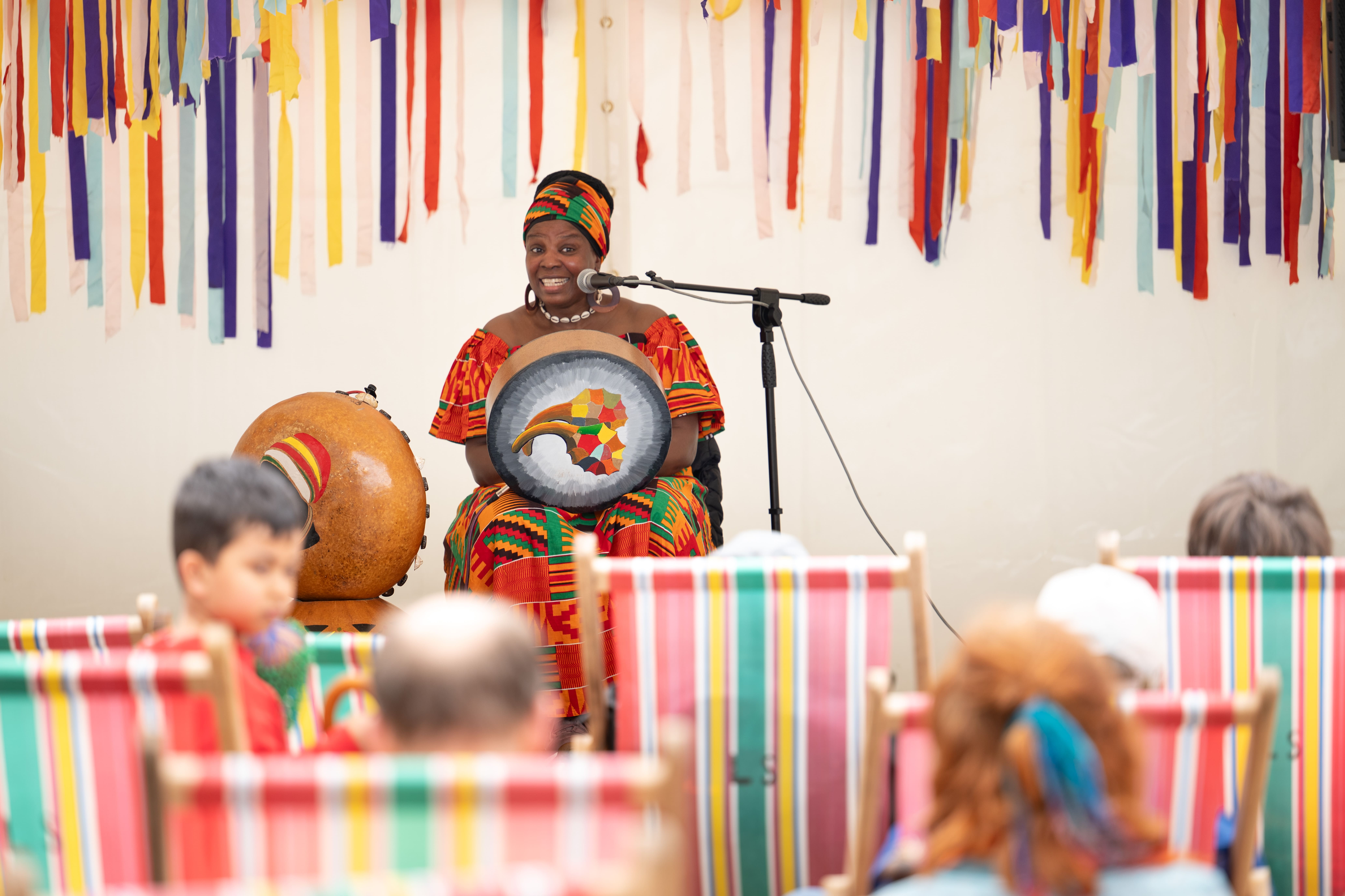A woman in colourful traditional attire sits on stage with a drum, smiling and speaking into a microphone. Bright striped decorations and chairs fill the scene, with an audience watching her performance.