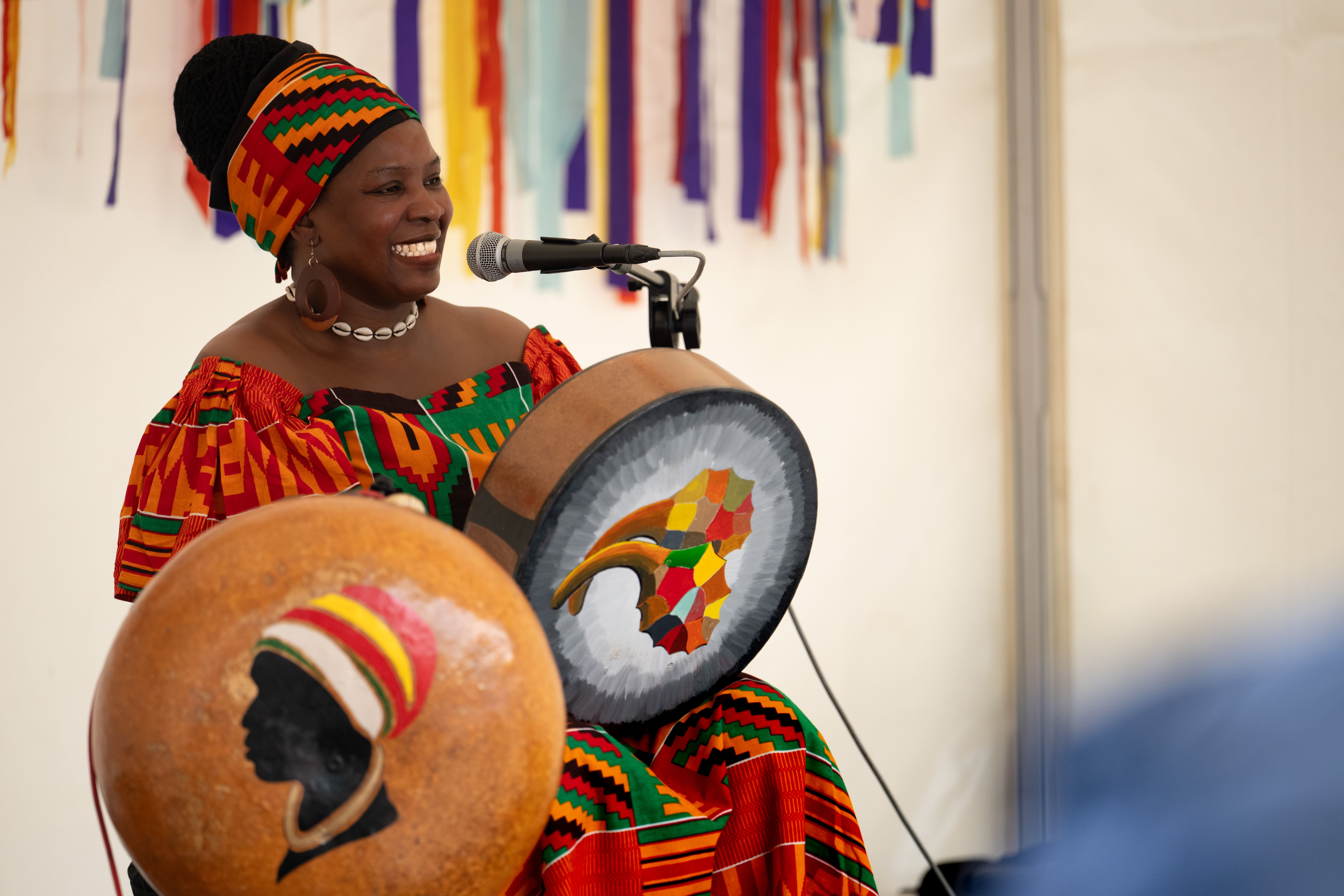 A smiling woman in colourful traditional African attire plays a painted drum and sings into a microphone, with multicoloured ribbons hanging in the background.