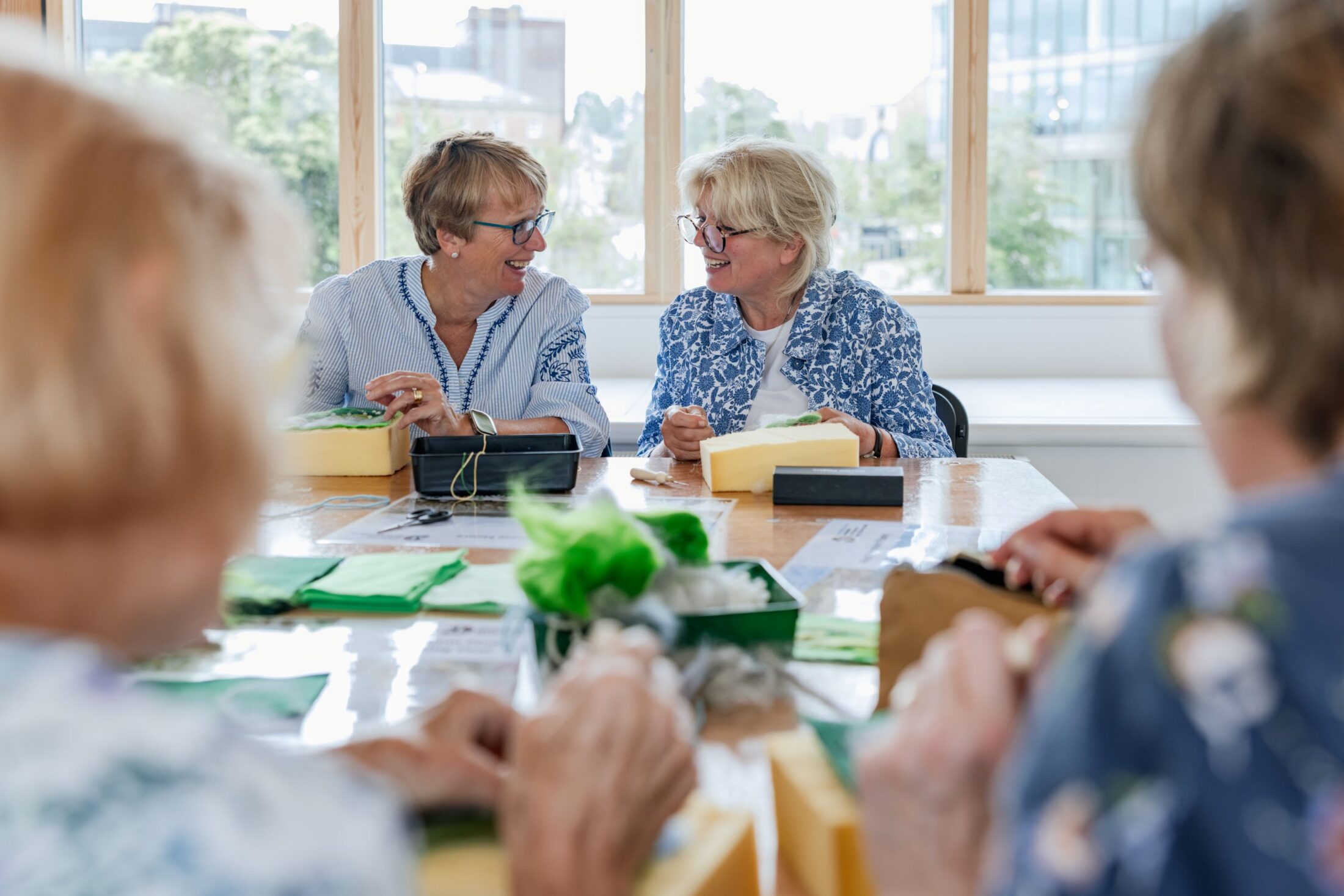 Two older women sit at a table, smiling and crafting together with green and yellow materials. Blurred people are in the foreground, and large windows show a cityscape outside.
