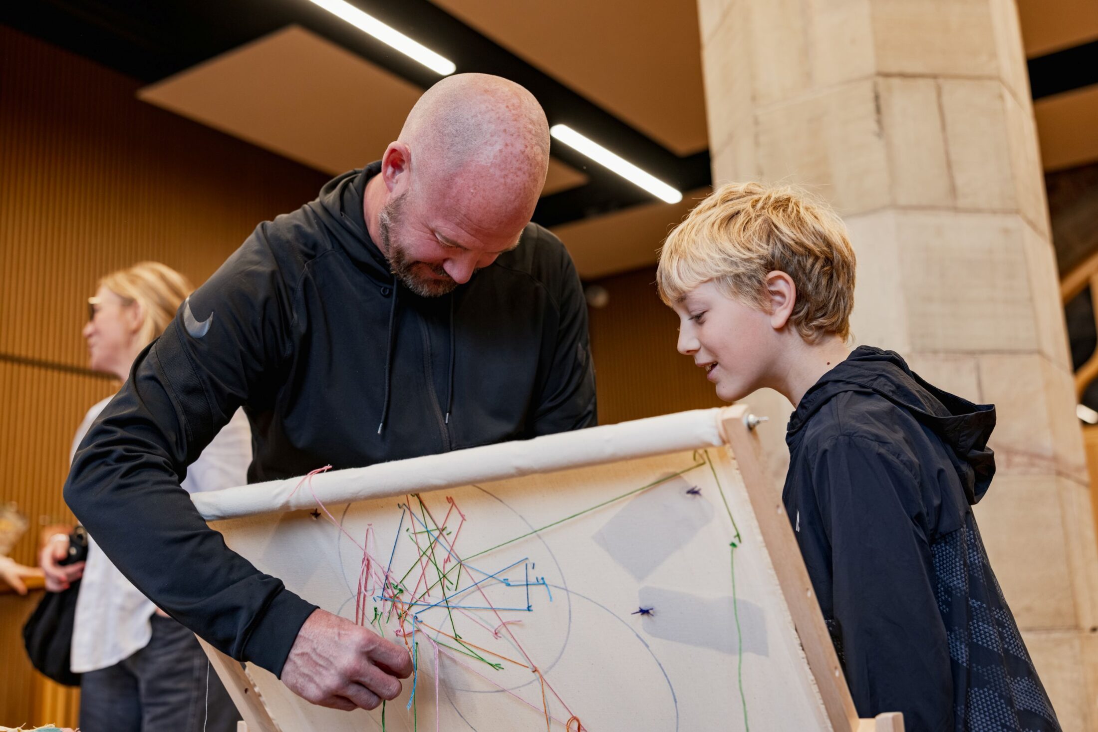 A man and a boy work together on a string art project, wrapping colourful threads around pins on a canvas in an indoor setting.
