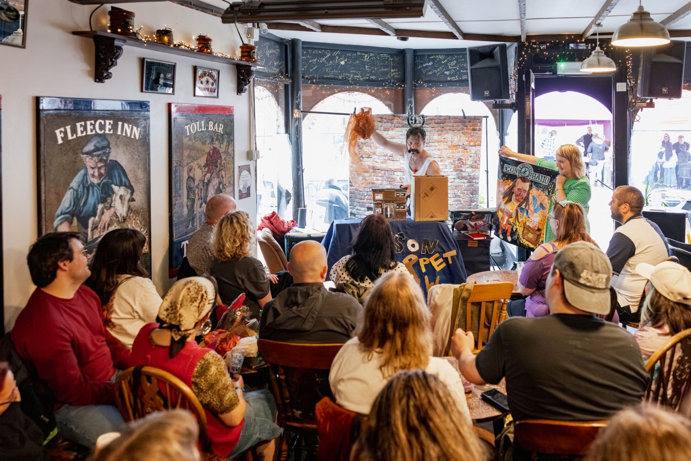 A performer stands at a table with a puppet, entertaining an audience seated in a cosy, art-filled café. Two people hold a colourful puppet show sign near the front.