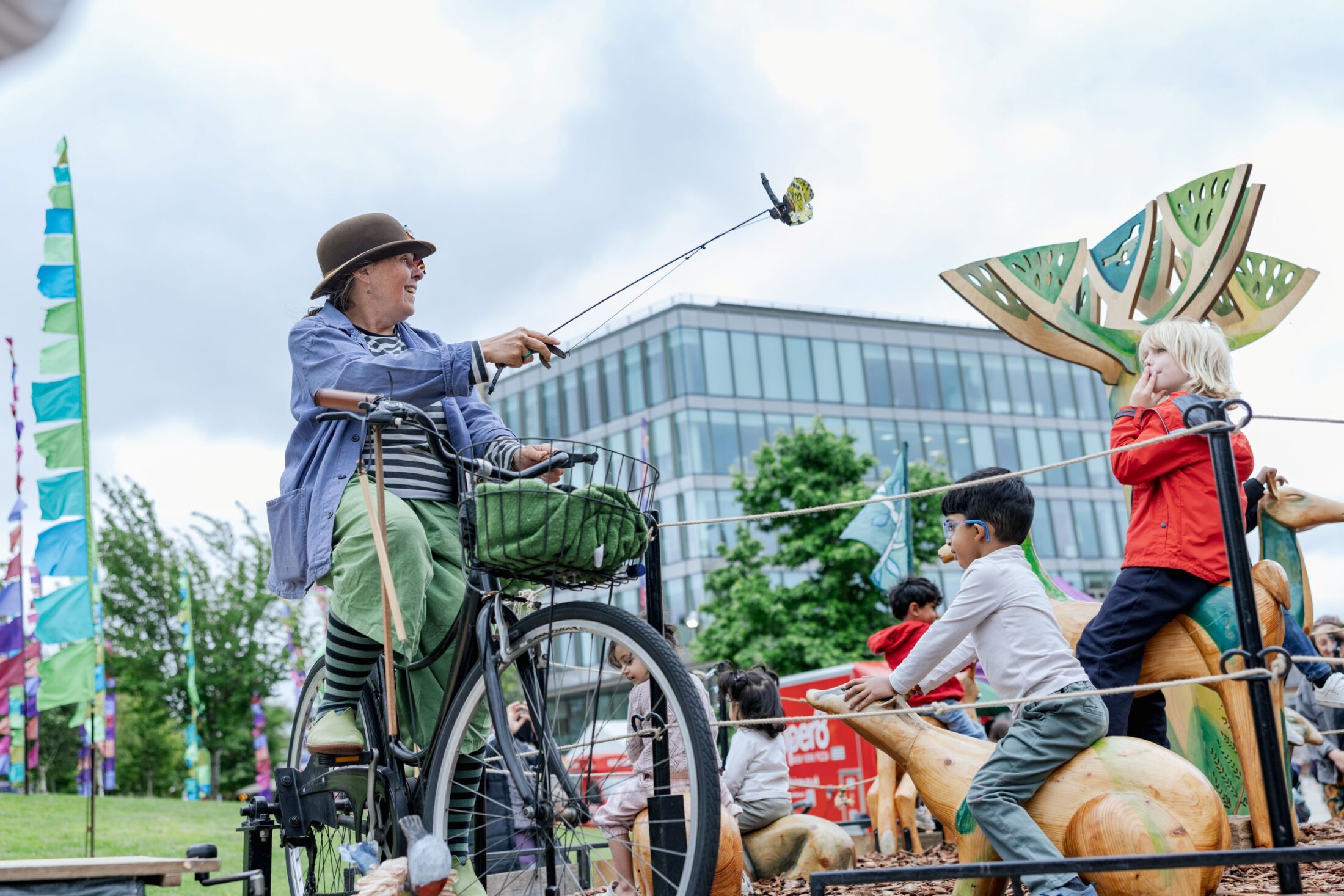 A woman on a bicycle entertains children by holding a puppet on a stick. Kids watch and play on animal sculptures outdoors, with colourful flags and a modern building in the background.
