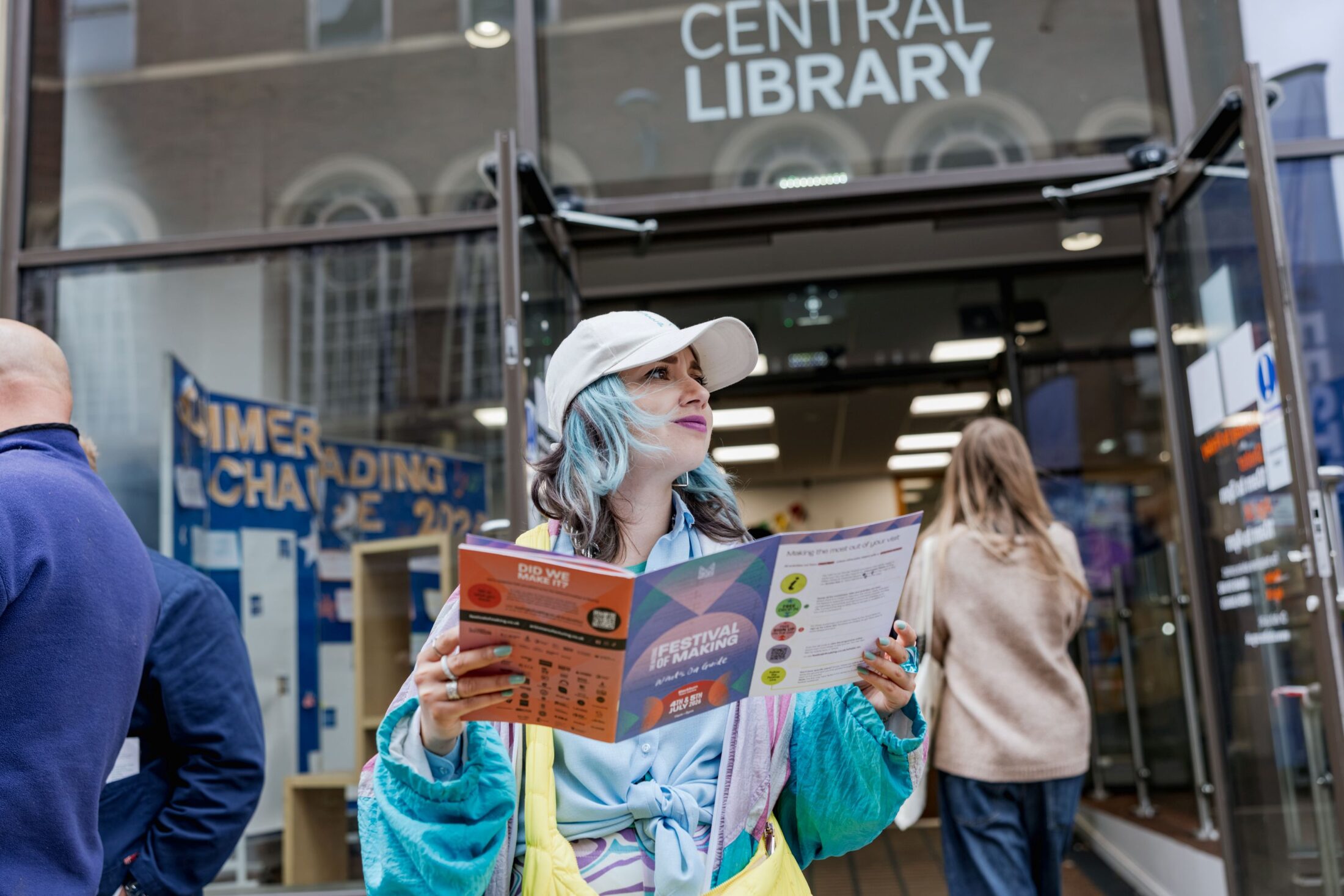 A person with dyed hair and a white cap holds a colourful event brochure while standing outside the entrance to a central library. Other people are nearby, and a SUMMER READING CHALLENGE sign is visible.
