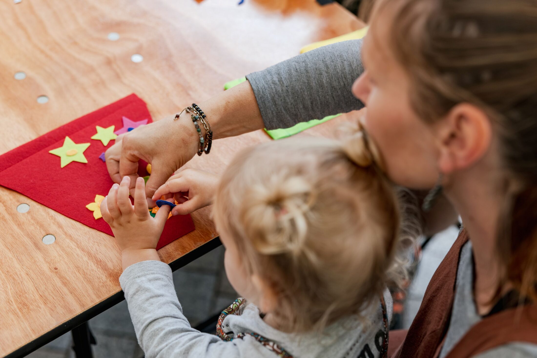 An adult helps a small child place colourful felt stars on a red piece of felt whilst sitting at a wooden table.