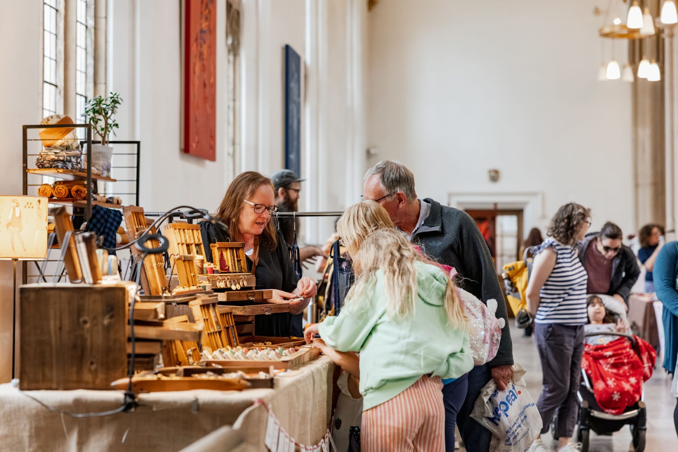 People browse wooden crafts at an indoor market stall. A woman stands behind the table helping customers, whilst others walk and shop in a bright, spacious hall with tall windows.