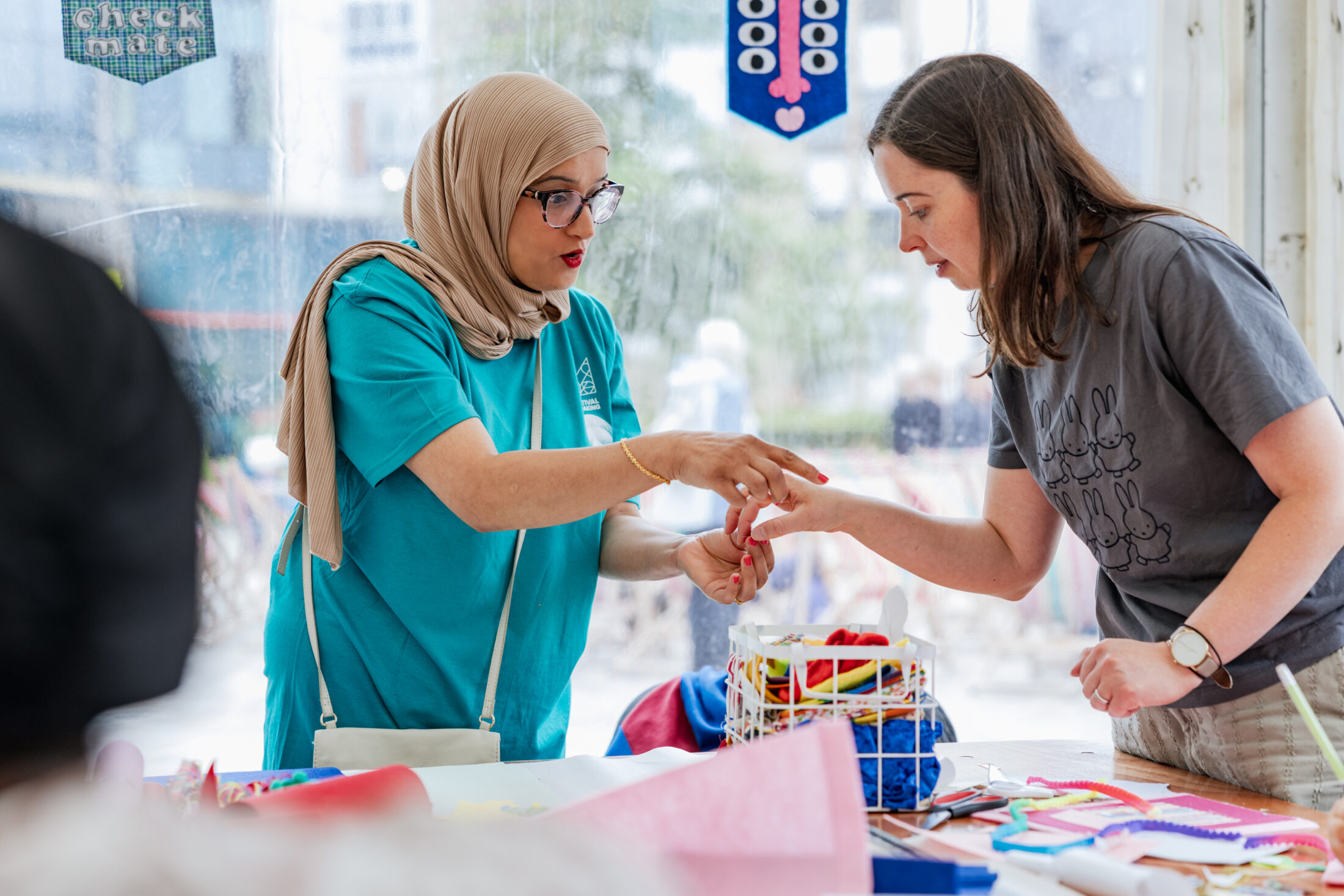 A woman in a beige hijab, wearing a light blue volunteer t-shirt assists a woman suring a felt banner making workshops in a marquee.