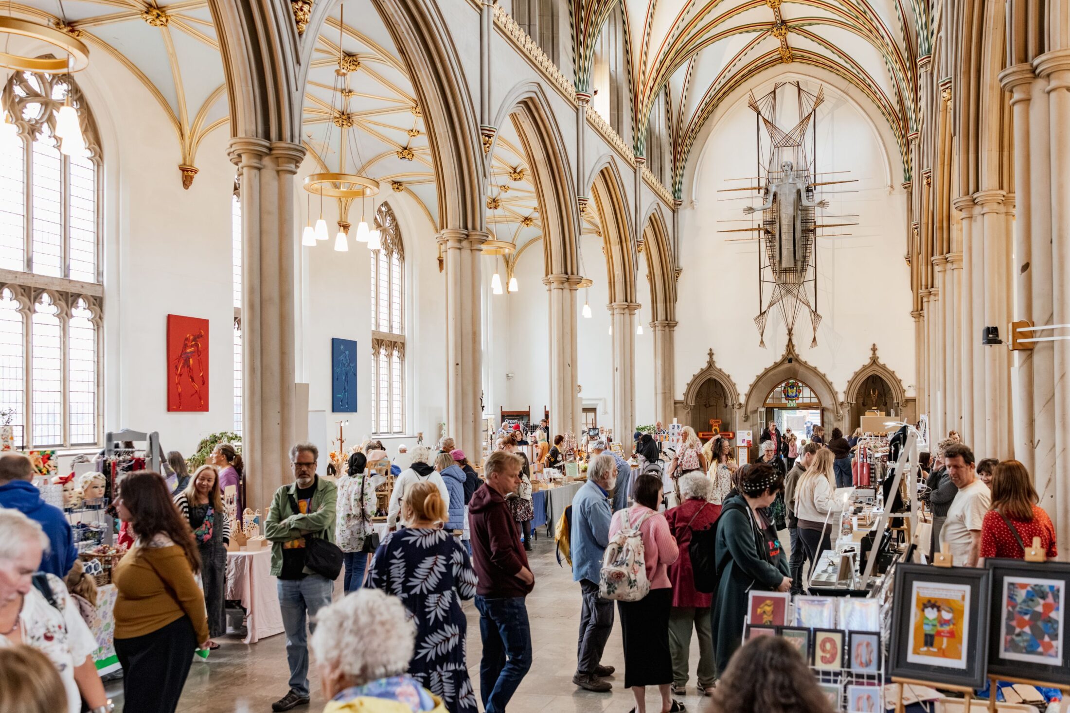 People browse various vendor tables inside a large, ornate hall with tall arched windows, columns, and vaulted ceilings. The space is bright and busy, filled with art and craft displays.