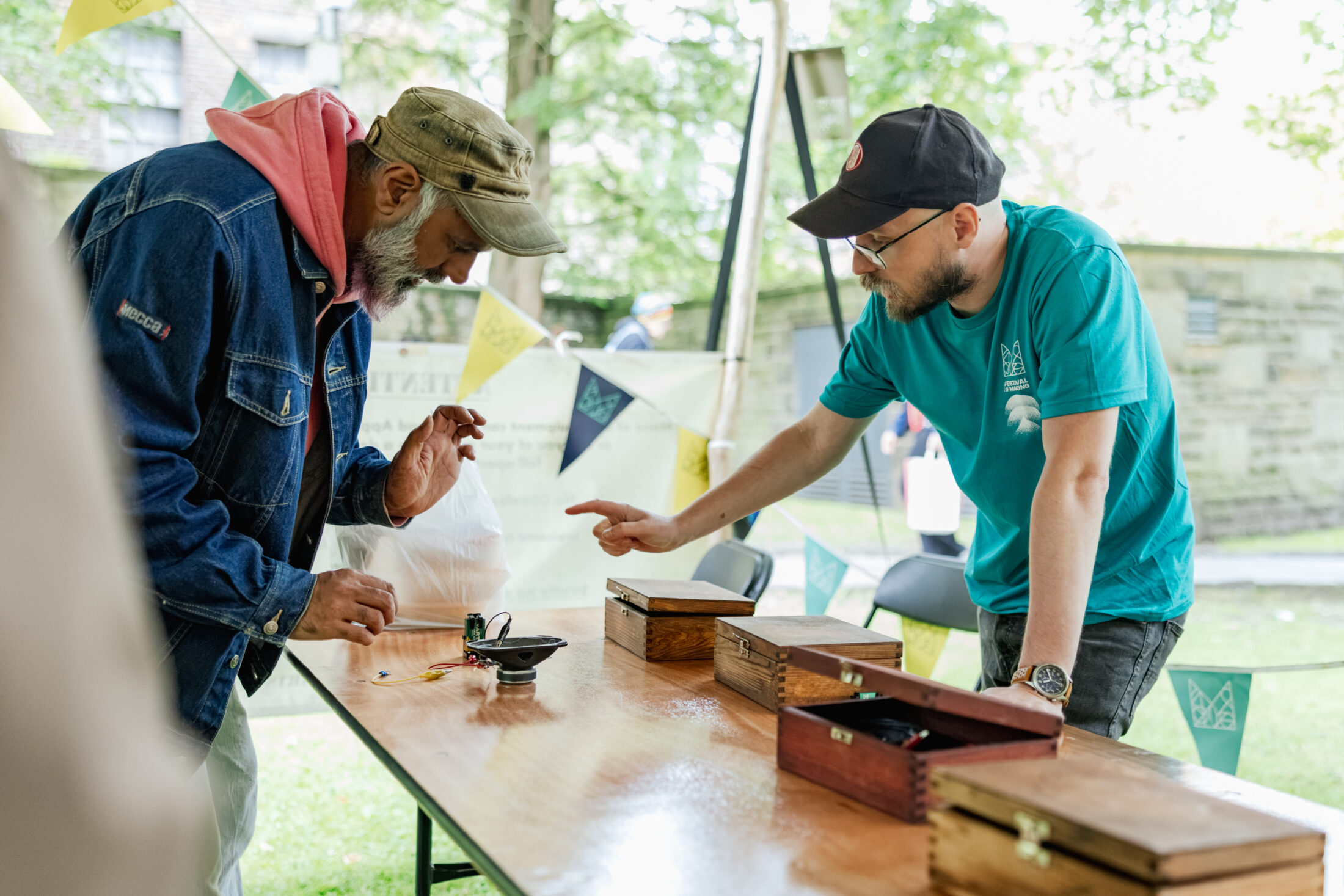 A man in a cap wearing a light blue volunteer t-shirt assists a man during a workshops under an open sided outdoor canopy.