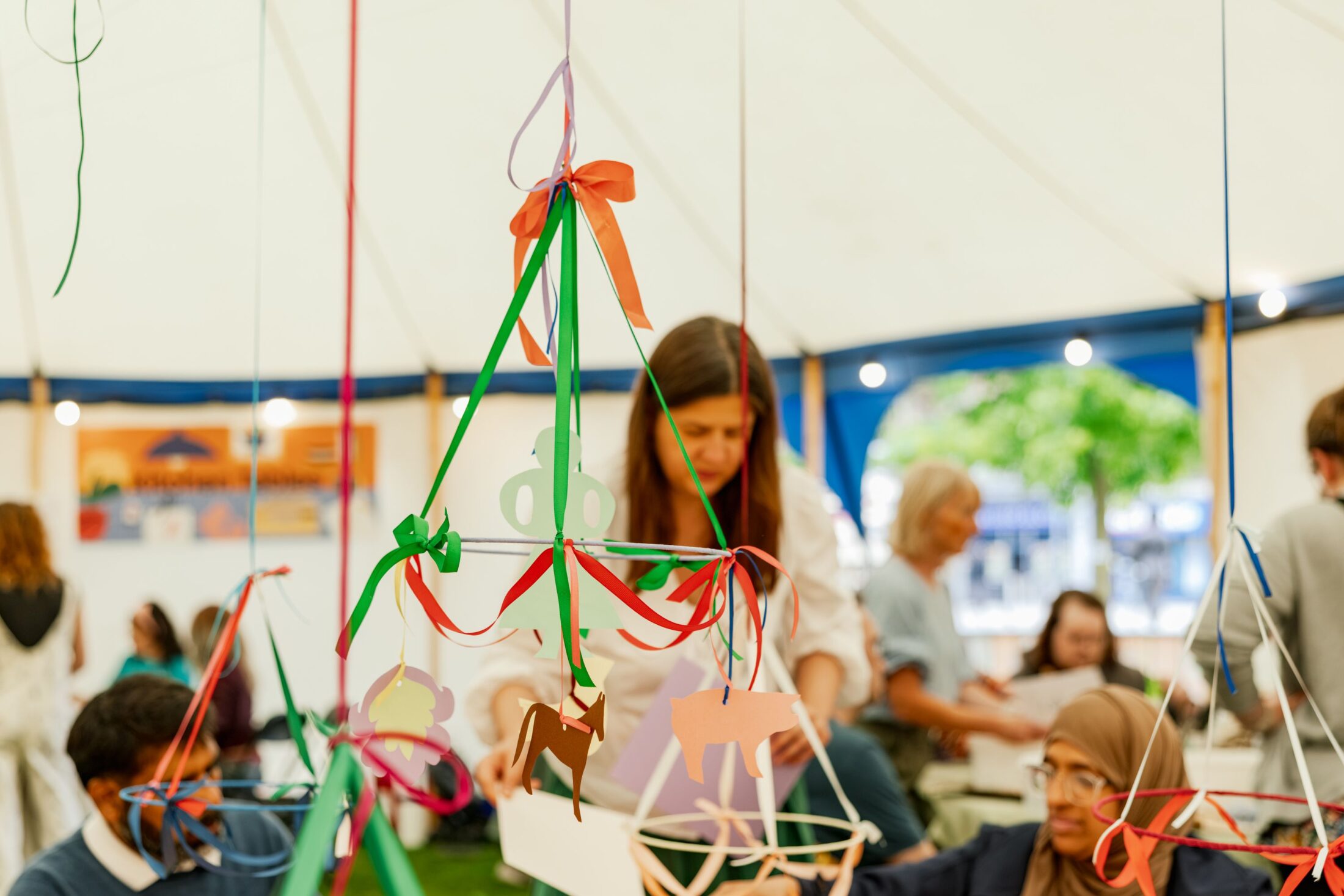 People in a tent work on a colourful mobile craft made of paper shapes and ribbons. A woman in the background focuses on the project, whilst others sit and create at tables.