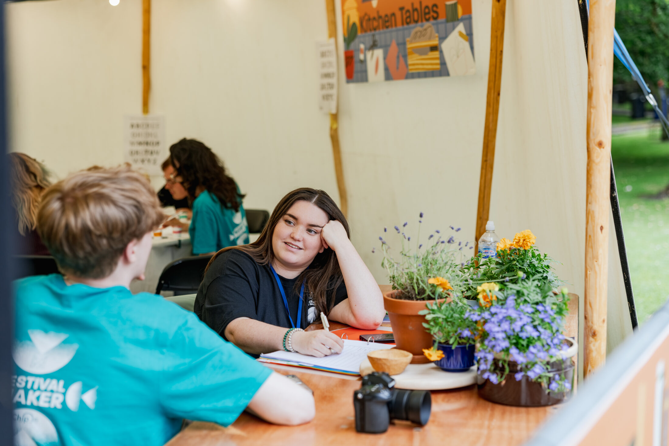 A woman sits at a table with a notebook, talking to a young person. Flowers and a camera are on the table. Other people work in the background inside a marquee at an outdoor event.