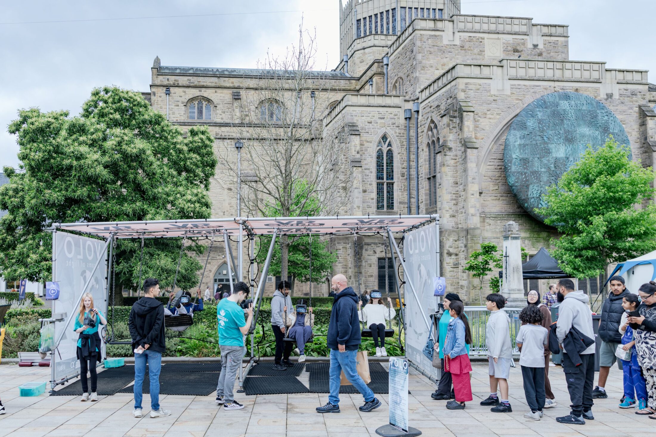 People queue outside a modern marquee, set up in front of a historic stone church. Some chat, while others wait, and trees and greenery surround the scene on a cloudy day.