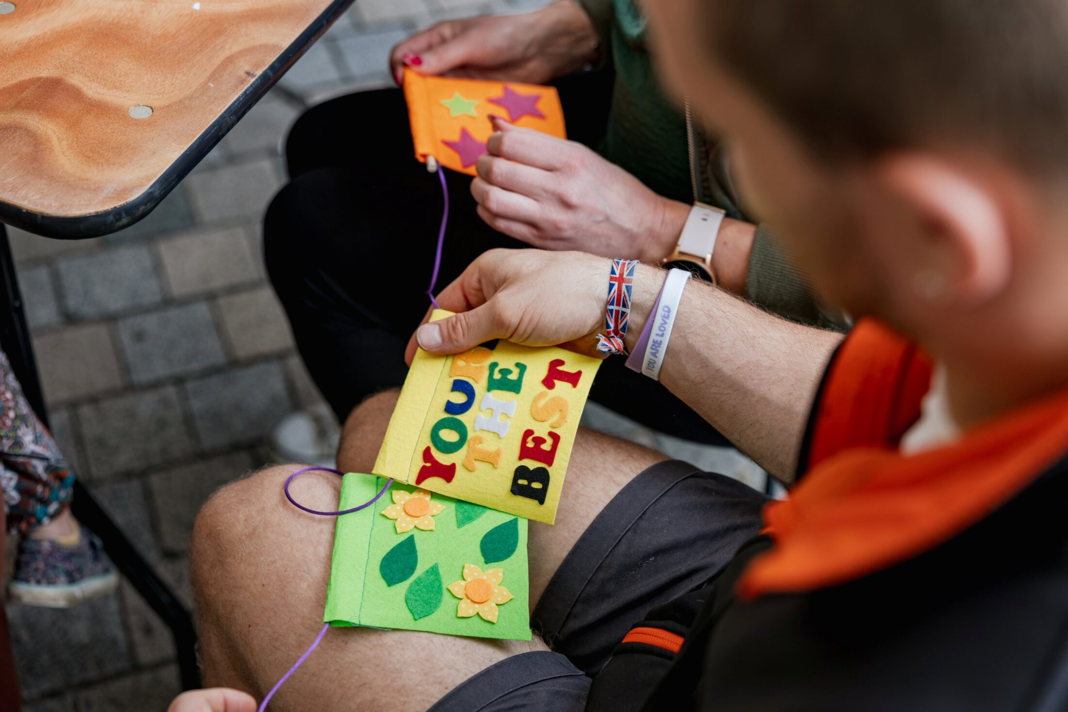 Two people sitting at a table work on colourful felt crafts. One holds a piece that reads YOU THE BEST decorated with flowers and leaves, whilst the other is crafting with stars.