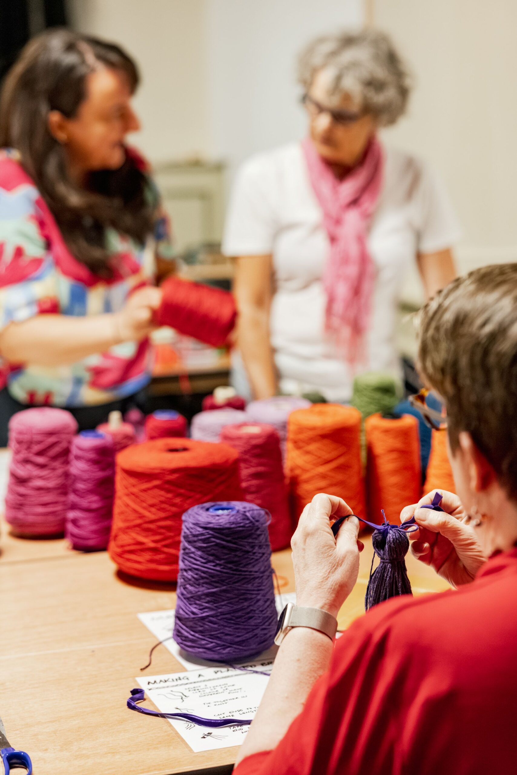 Three people gather around a table covered with colourful yarn cones. One person in the foreground works with purple yarn, whilst the others chat in the background. The focus is on crafts and creativity.