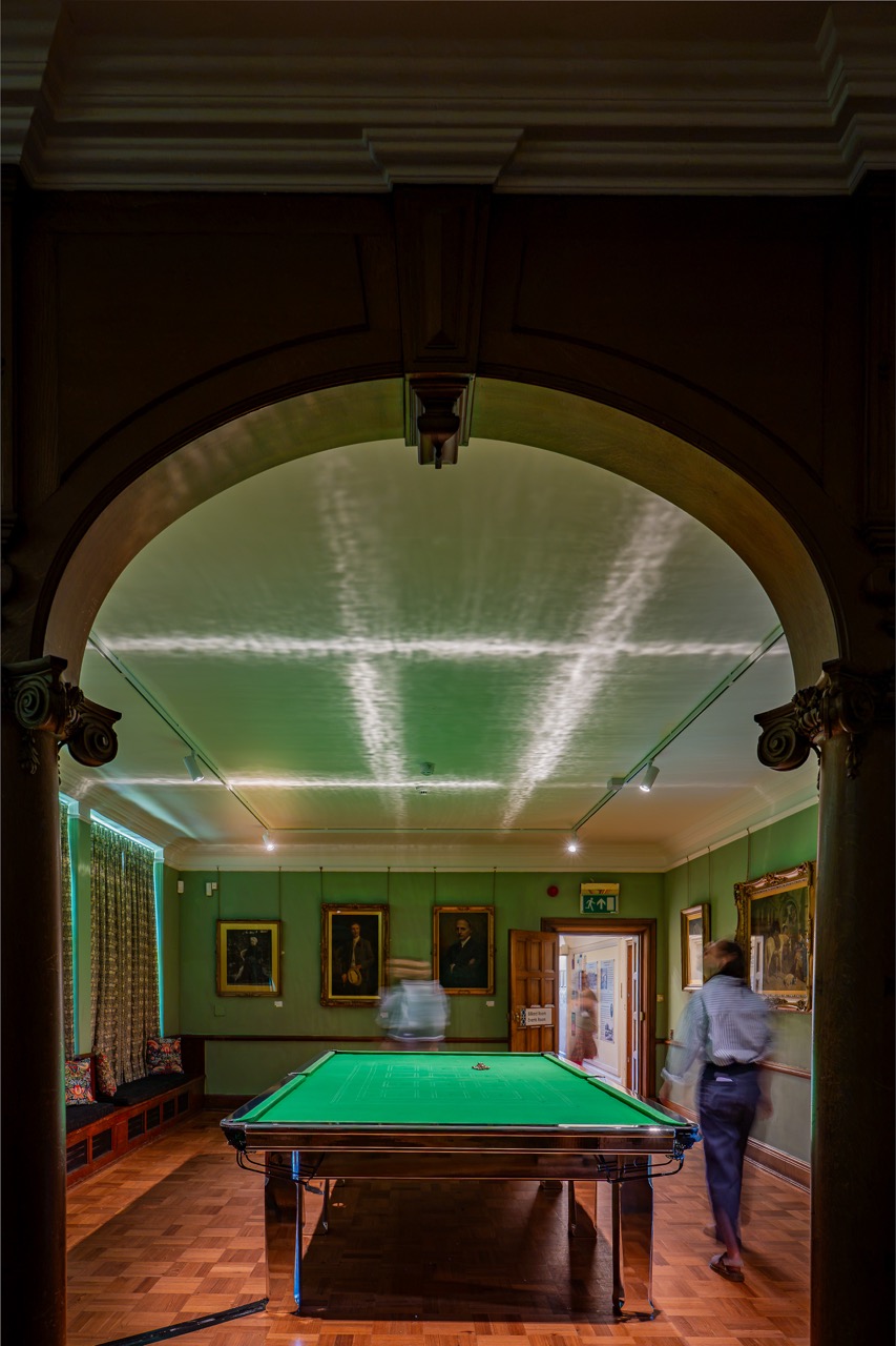 A snooker table is centred under an archway in a warmly lit room, with blurred figures near the table and paintings on the walls. Light reflections create patterns on the ceiling.