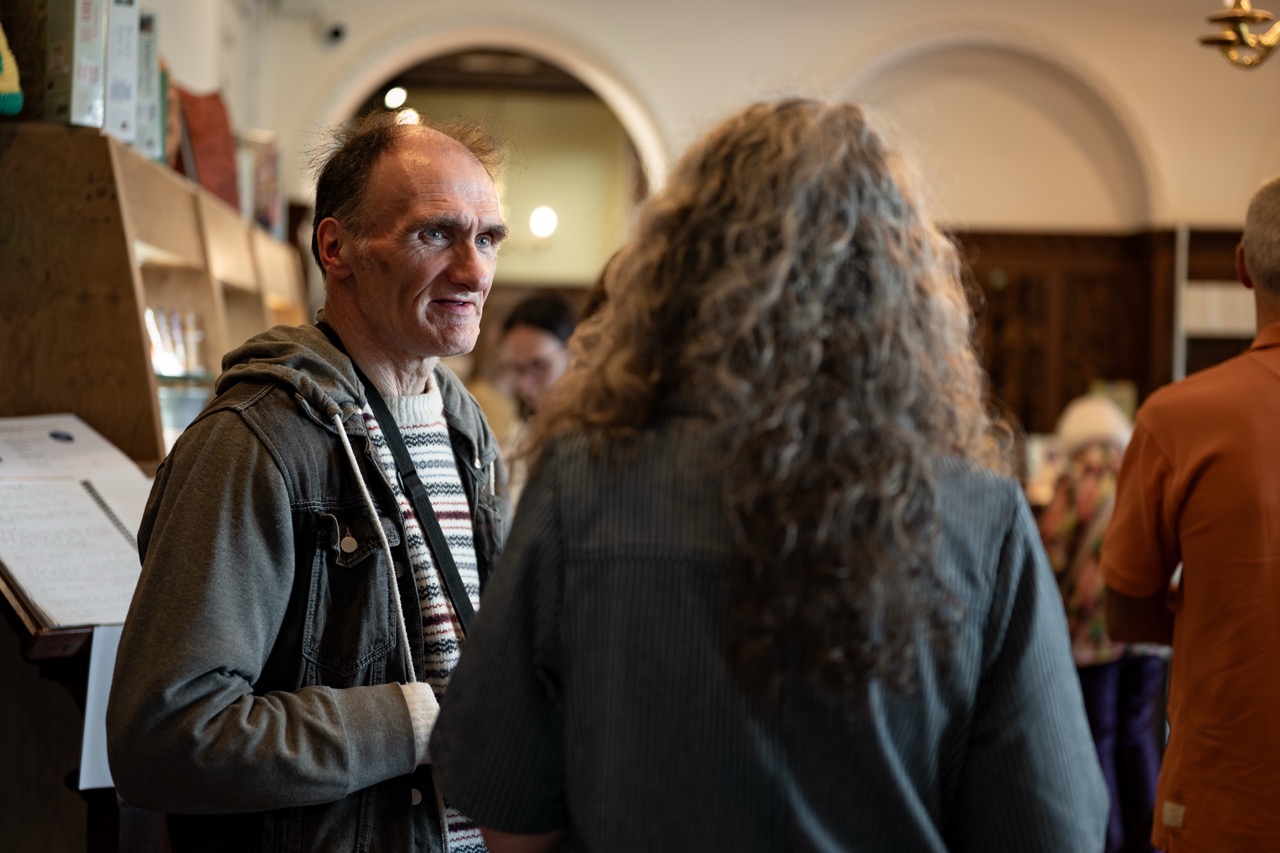 A man in a striped jumper and jacket talks to another person with long curly hair in a warmly lit room with people in the background.