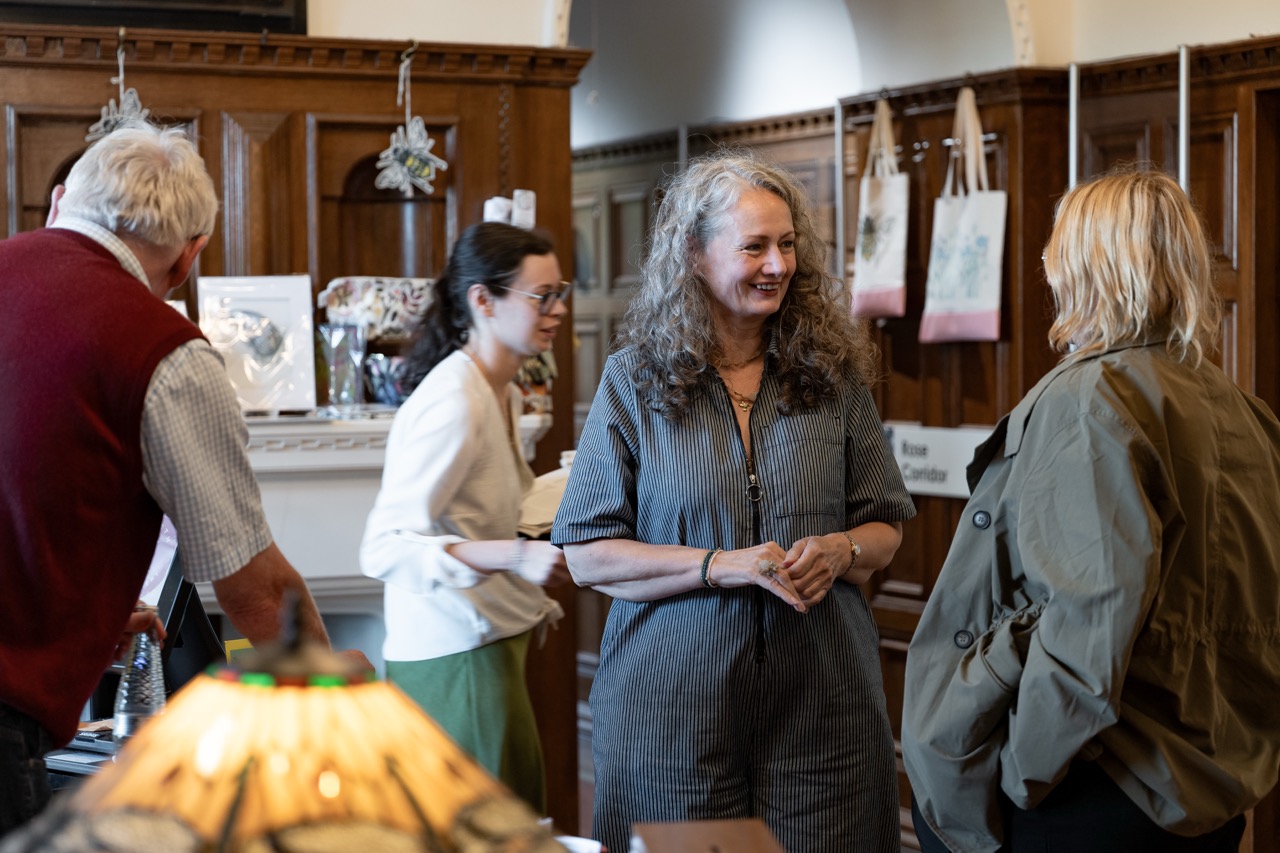 Four adults are talking and smiling in a warmly lit room with wood panelling and art displays. Two women are in focus, one with long grey hair, while two others are slightly blurred in the background.