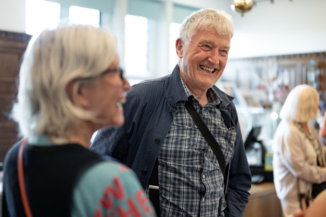 An older man and woman smile and talk in a brightly lit room, with blurred people and shelves in the background.