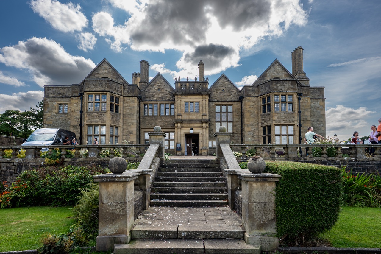 A grand stone mansion with tall chimneys and gabled roofs, seen from a staircase leading up to the entrance, surrounded by manicured gardens and people on either side.