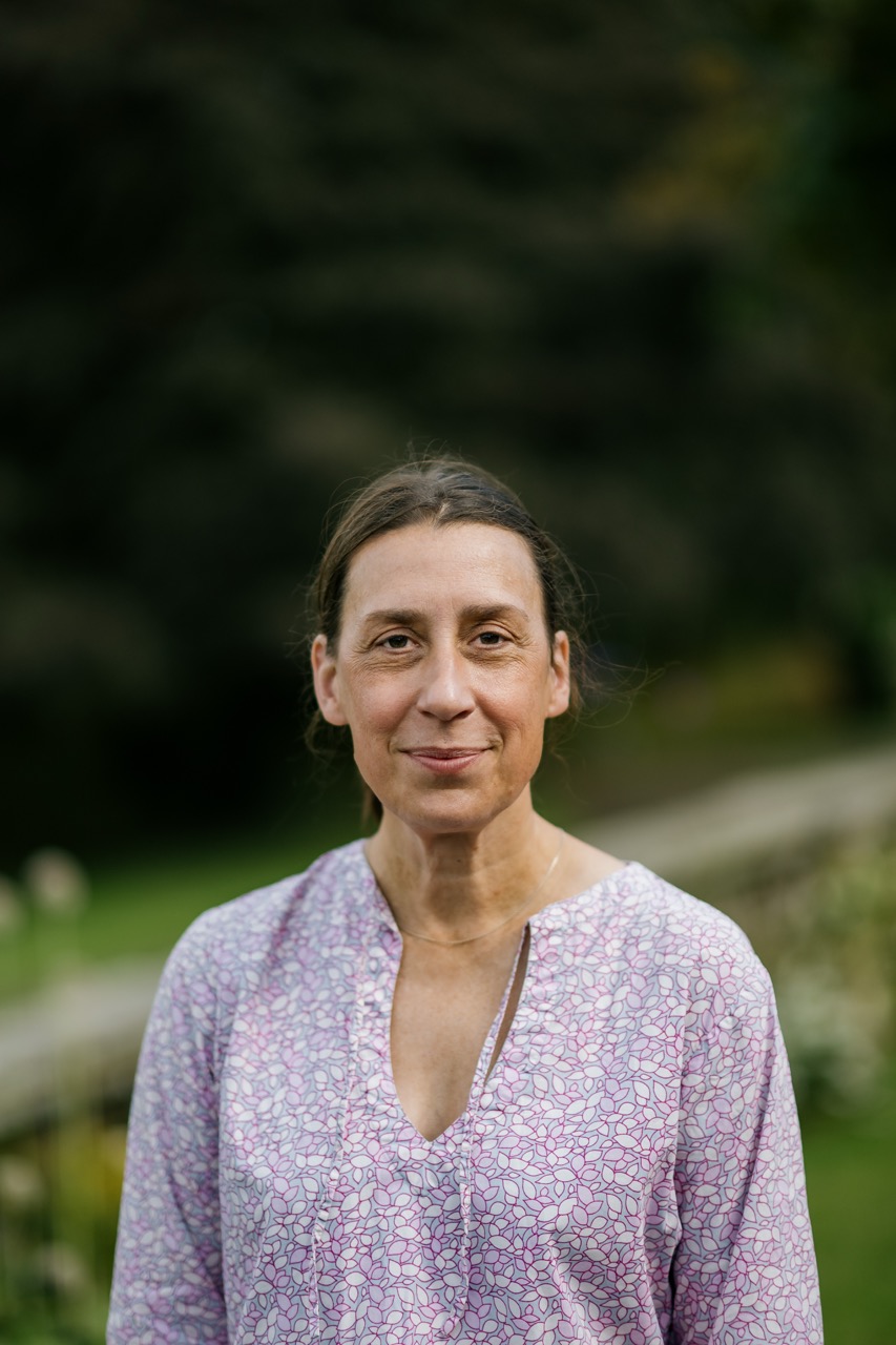 A person with long brown hair tied back, wearing a light purple patterned blouse, stands outdoors with greenery and a stone wall in the blurred background.