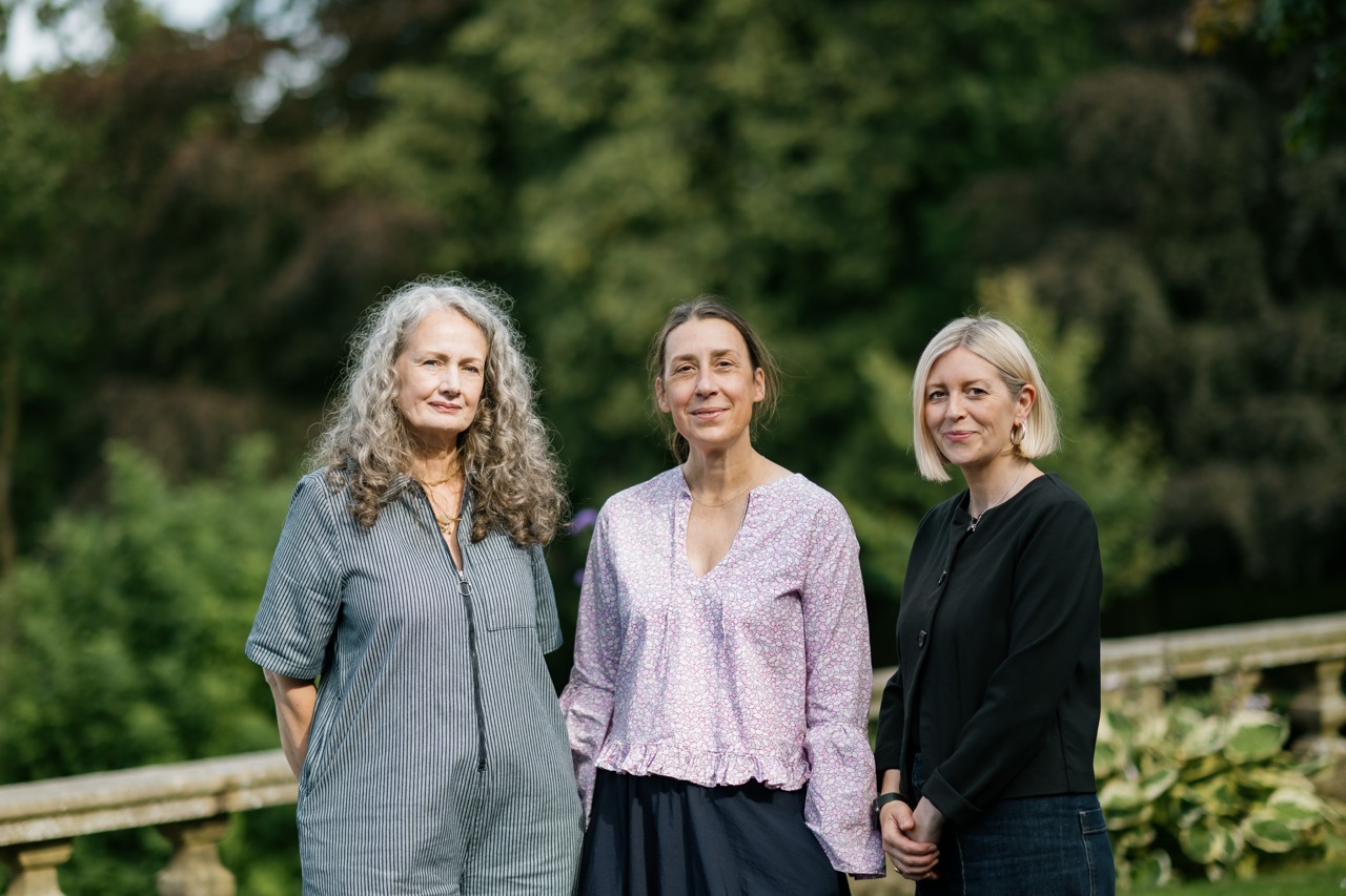 Three women stand outdoors in front of greenery, smiling softly at the camera. One has long curly grey hair, one has brown hair tied back, and one has short blonde hair.