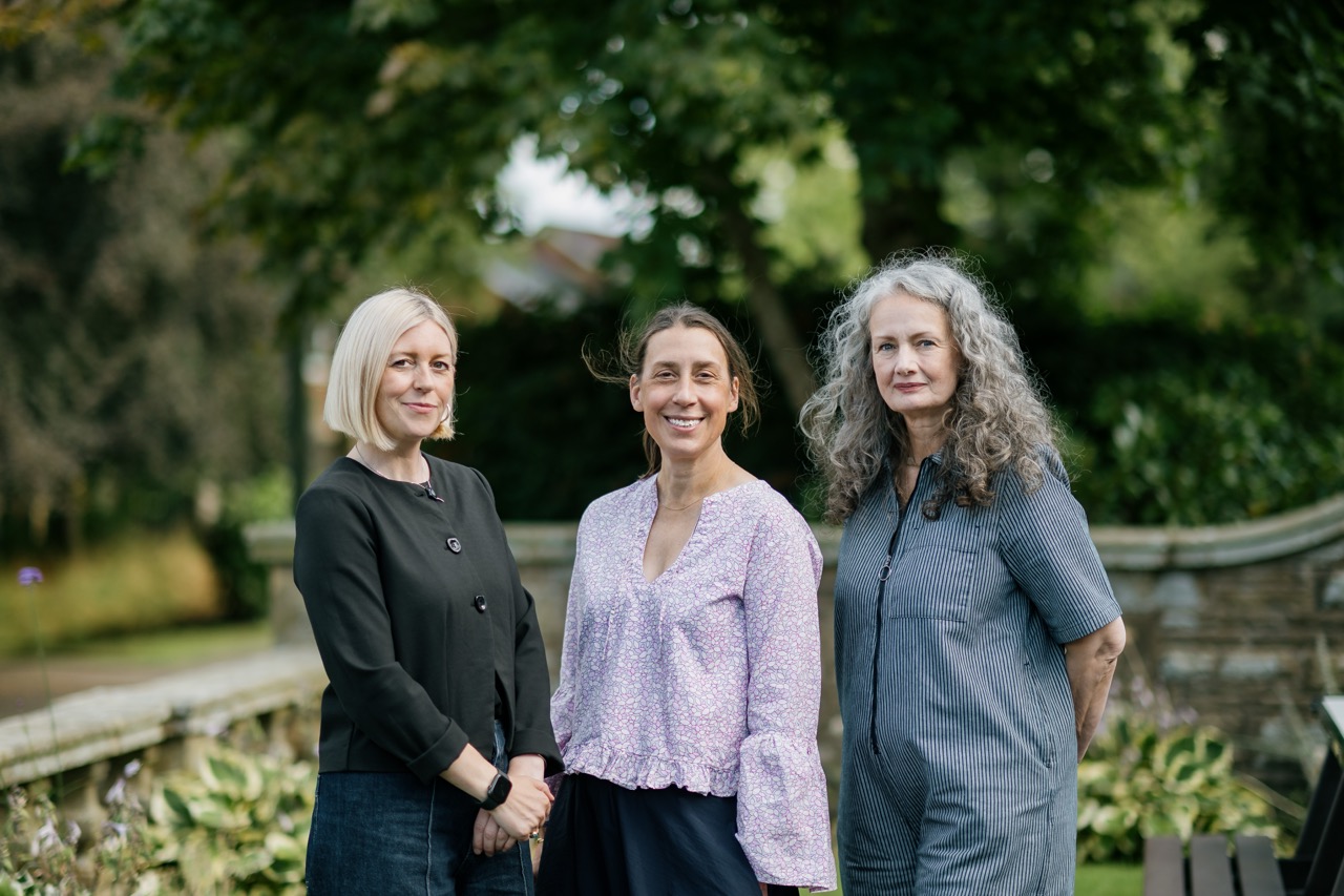 Three women stand outdoors, smiling, with greenery and a stone wall in the background. One wears a black jacket, one a light blouse, and one a grey dress.