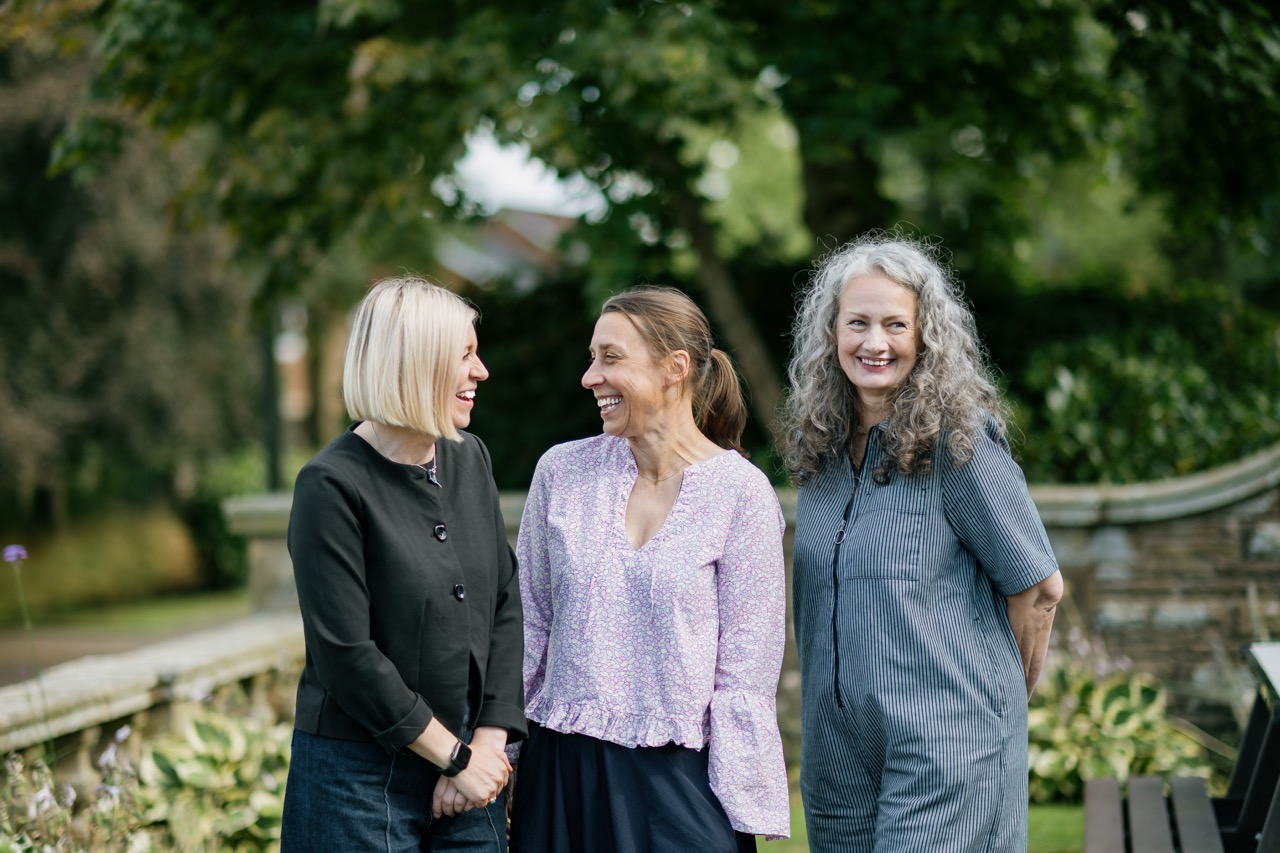 Three women stand outside, smiling and talking. Two have straight, fair hair, and one has curly, grey hair. They are casually dressed, with trees and greenery in the background.